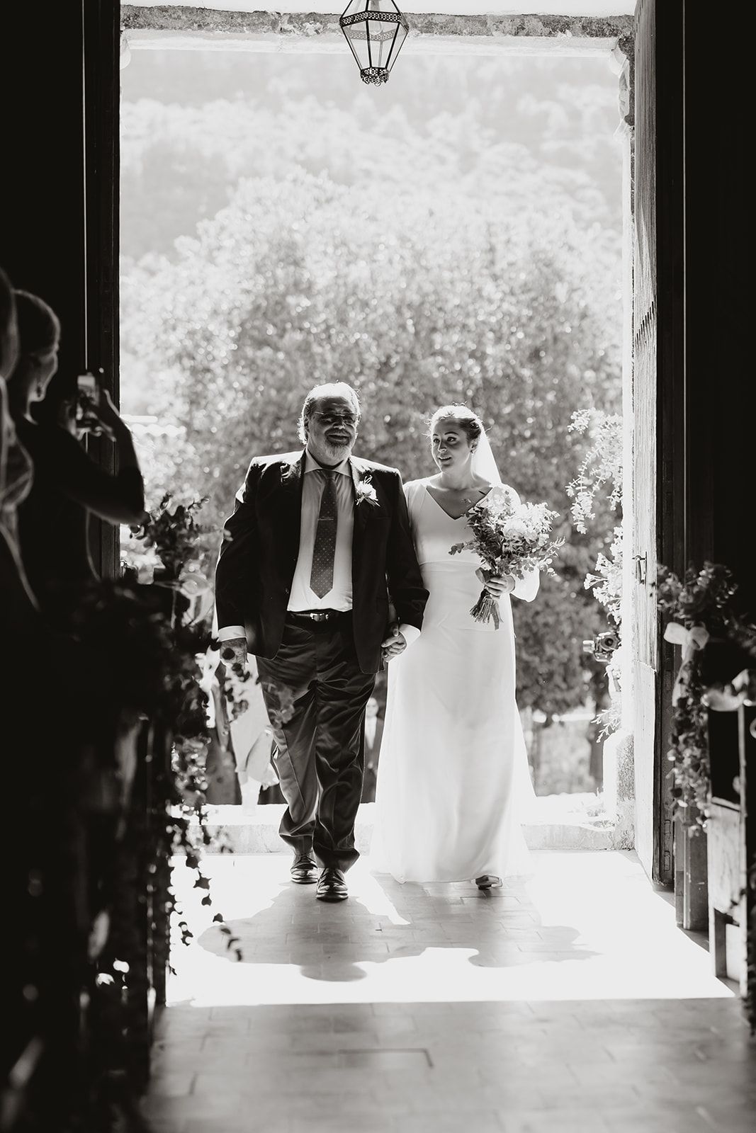 Black and white photo of wedding processional through sunlit church entrance.