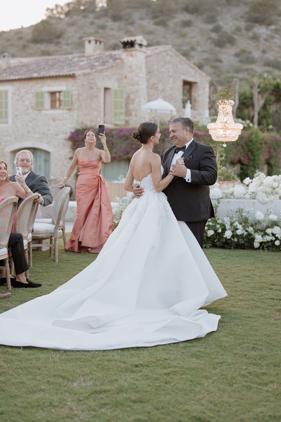 A wedding celebration on a lawn with a Mediterranean-style villa and hanging chandeliers in the background.