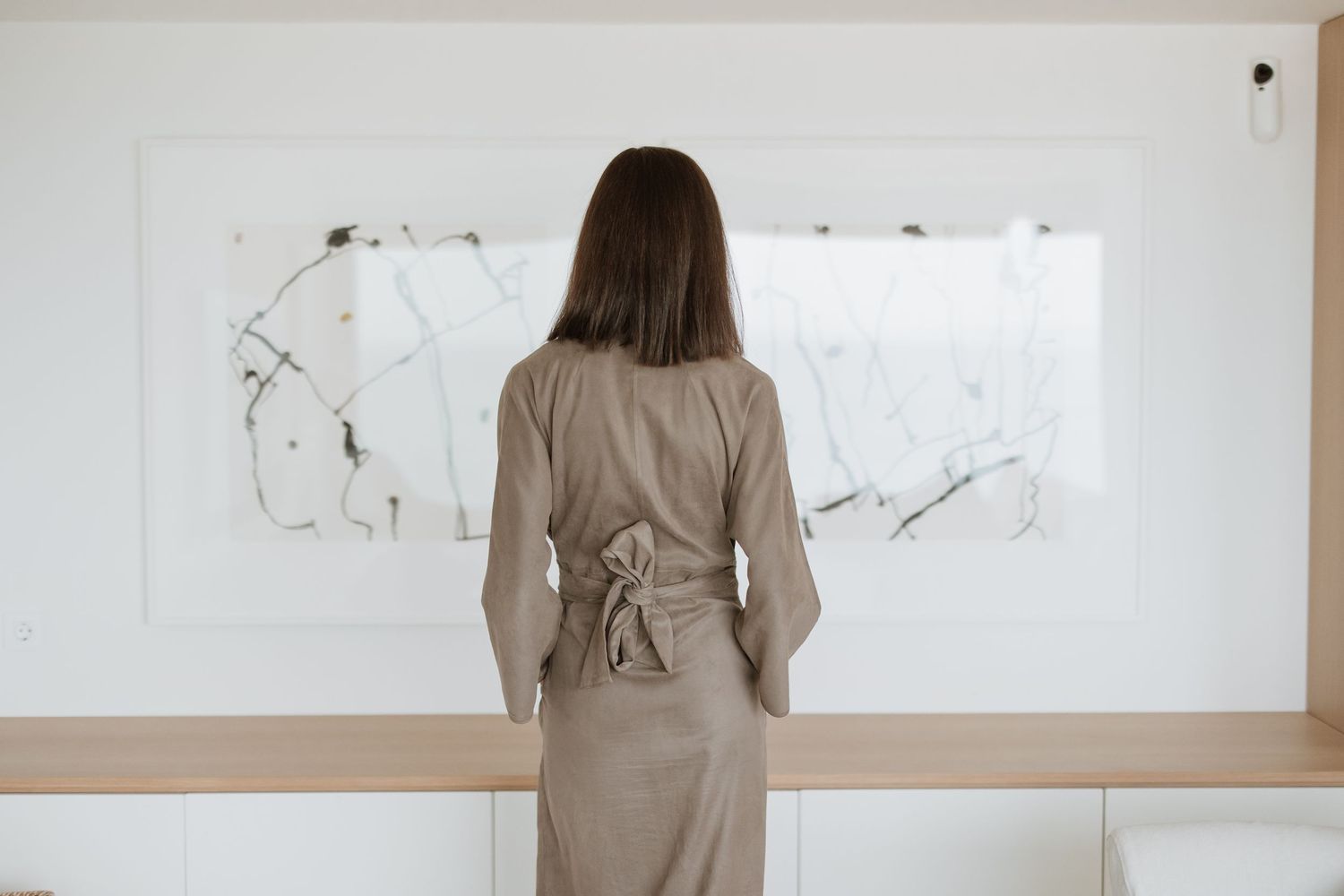 Back view of person in taupe dress standing before abstract white wall art.