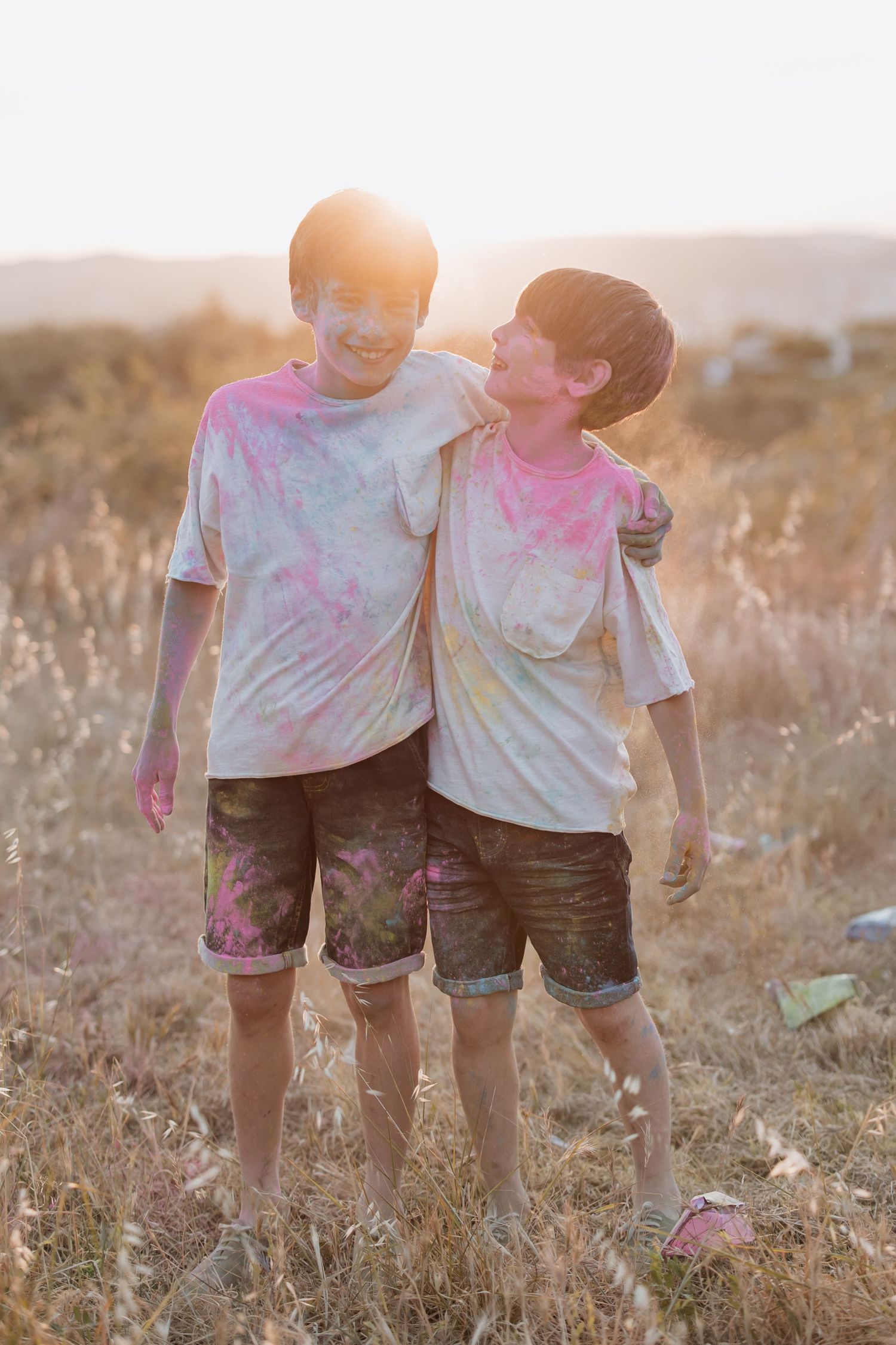 Two people wearing powder-covered white t-shirts and shorts stand together in a dry field at sunset after a color run event.