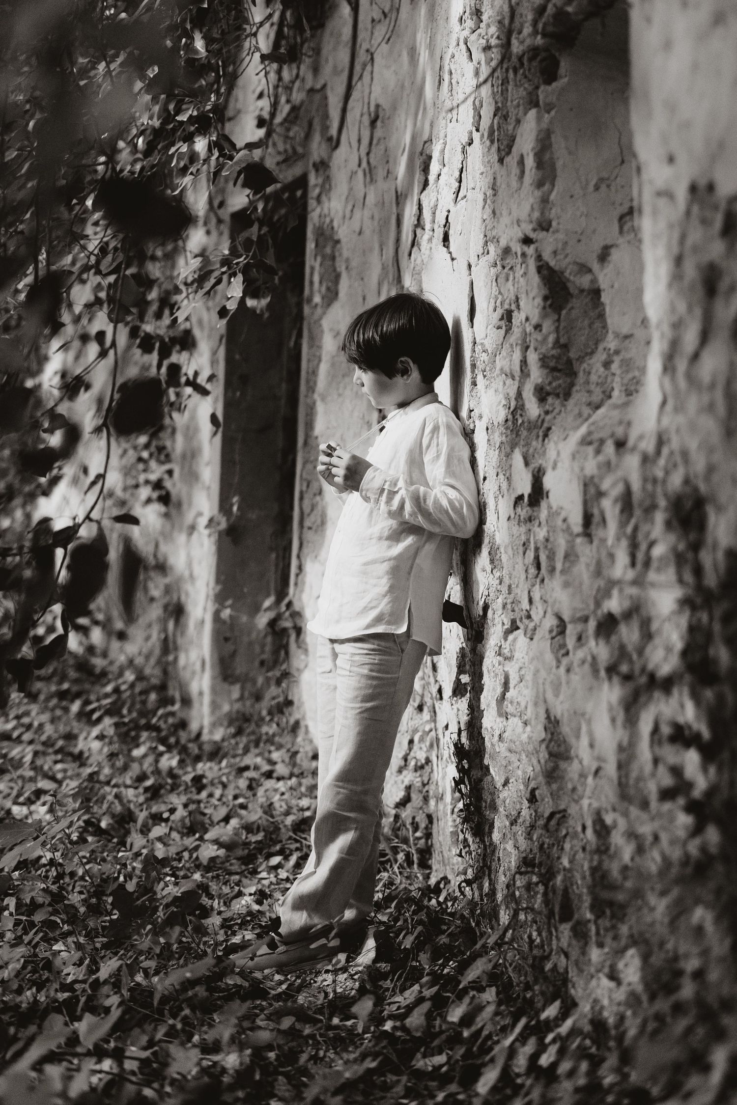 A black and white photograph shows a young figure in white clothing leaning against a weathered stone wall among autumn leaves.