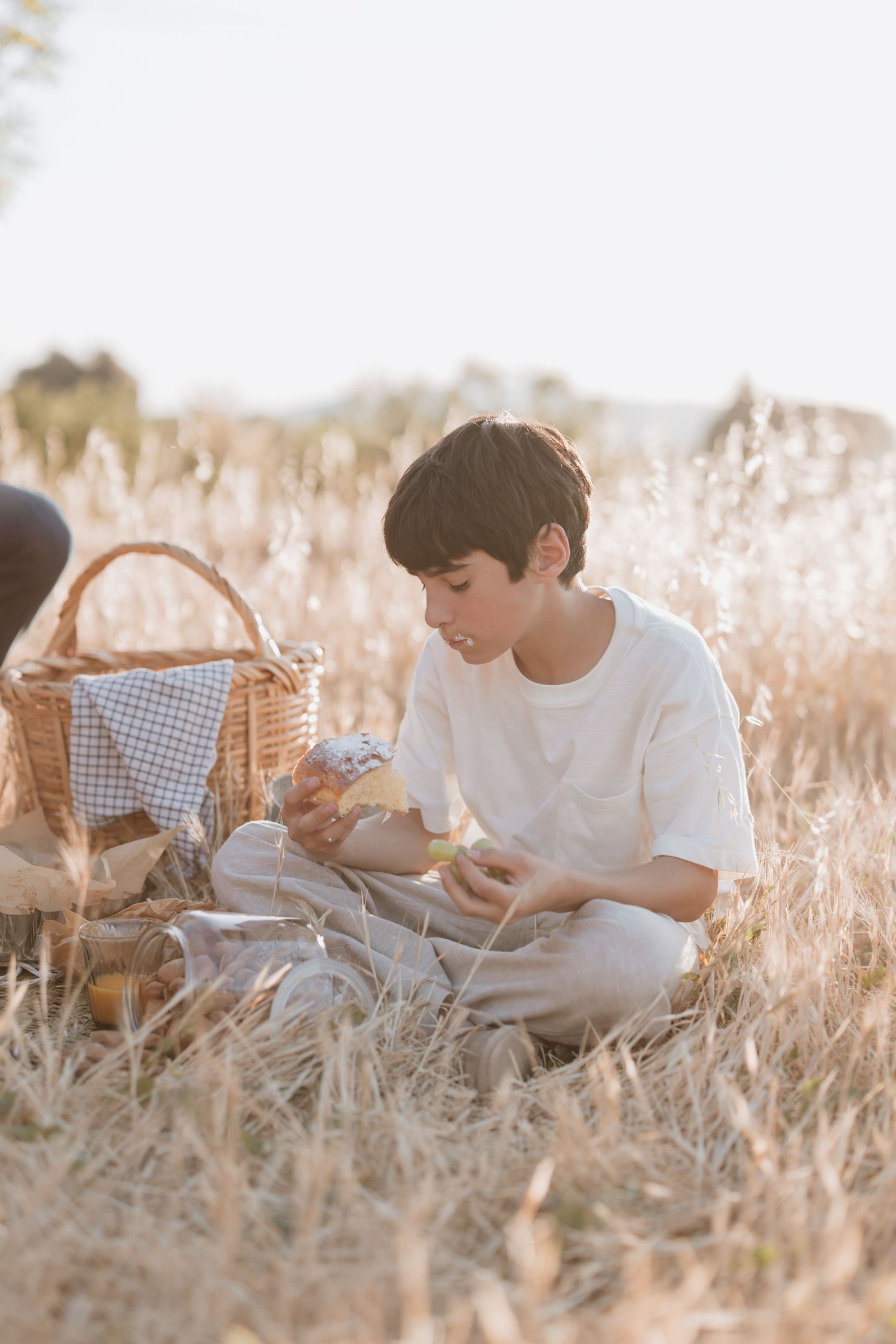 A picnic scene in a sunlit wheat field shows two people sitting together with a woven basket and food items.