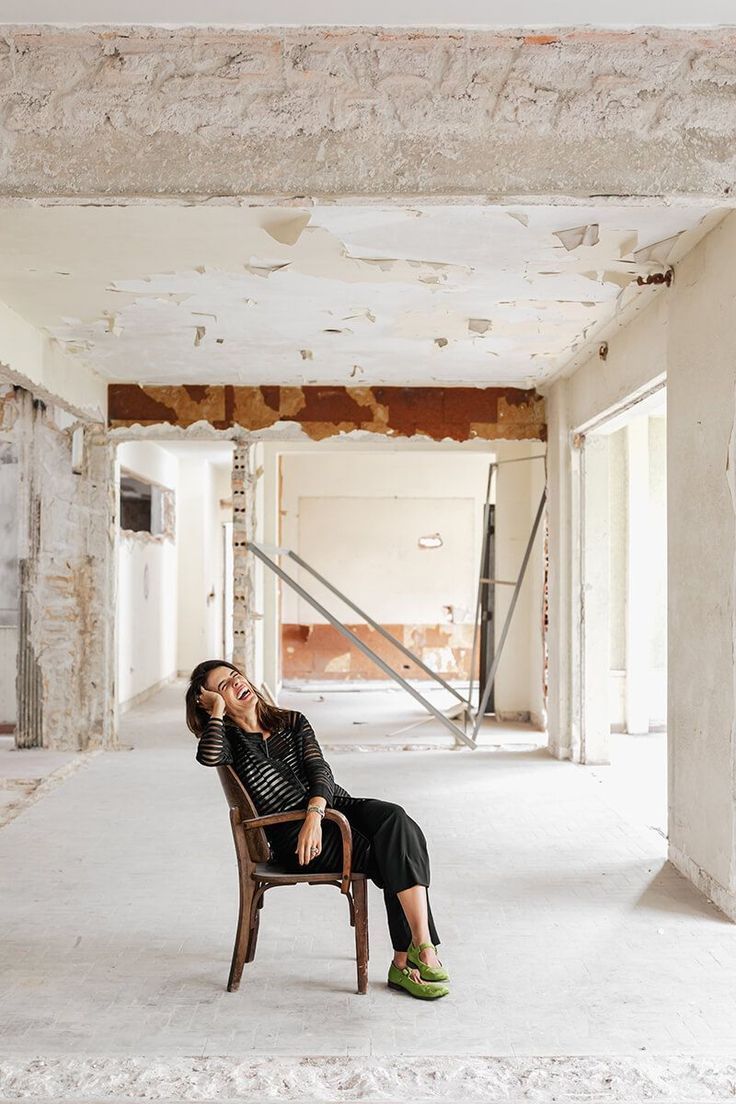 Woman in black outfit and green shoes sits on wooden chair in abandoned building with peeling walls and ceiling.