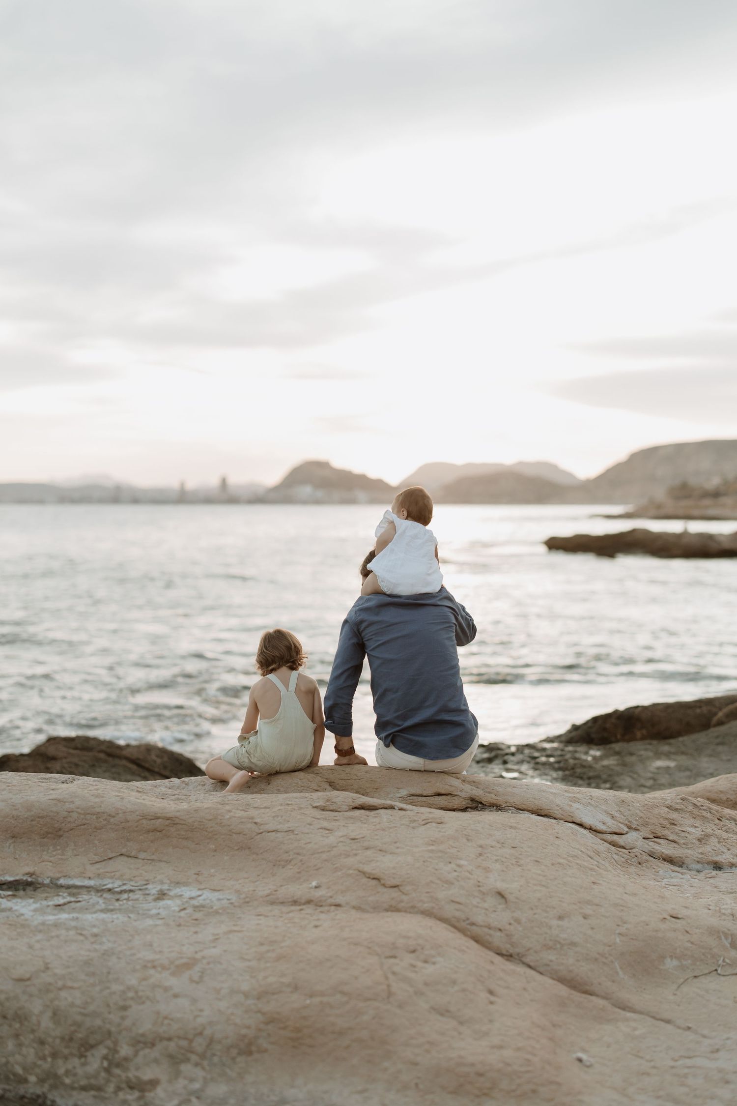People sitting on rocky shoreline watching the sunset over distant mountains.