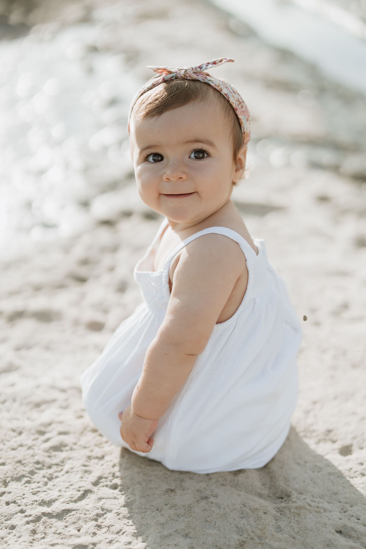 A baby in white dress smiles while playing on beach sand.