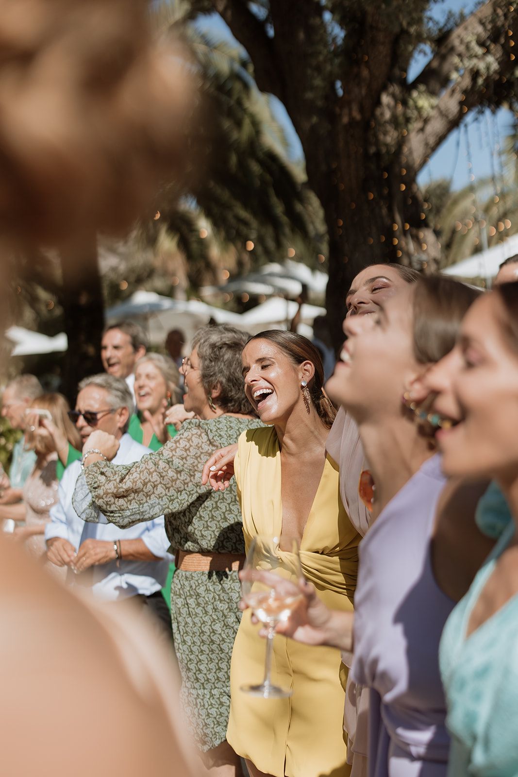 Wedding guests laugh together while holding drinks at an outdoor celebration.