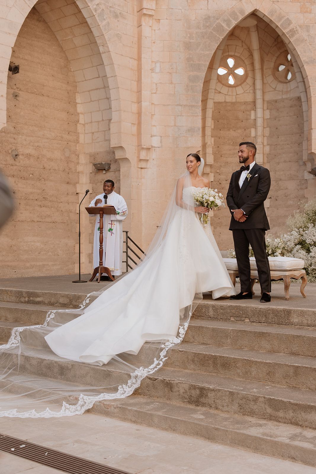 A wedding ceremony unfolds on stone steps with a dramatic cathedral veil trailing behind the couple.