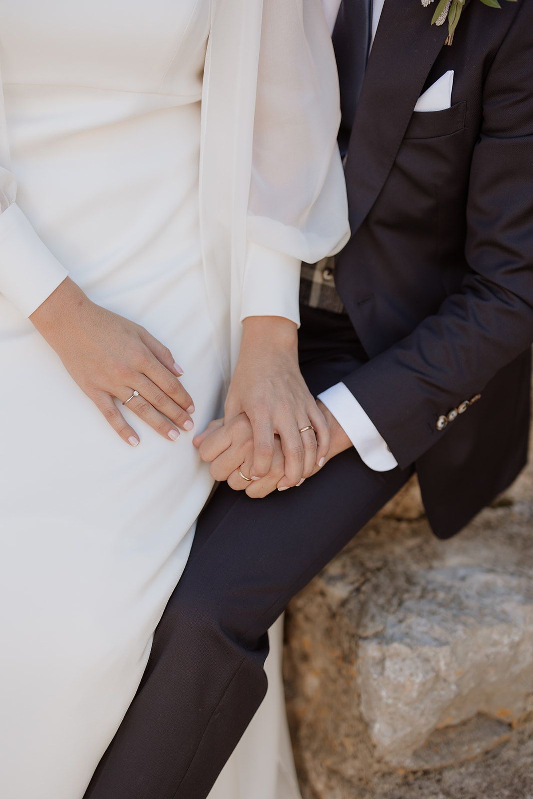 Close-up of newlyweds holding hands, showcasing wedding rings and formal attire.