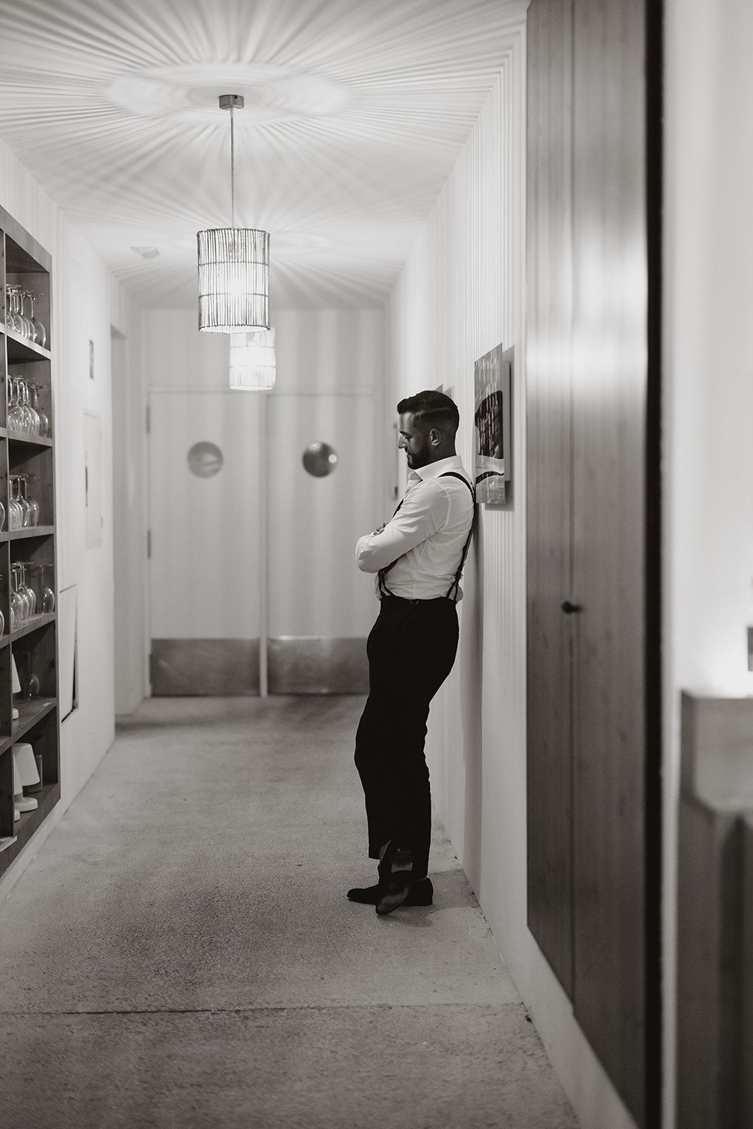 A person in formal attire stands contemplatively in a hallway with modern lighting and bookshelves.