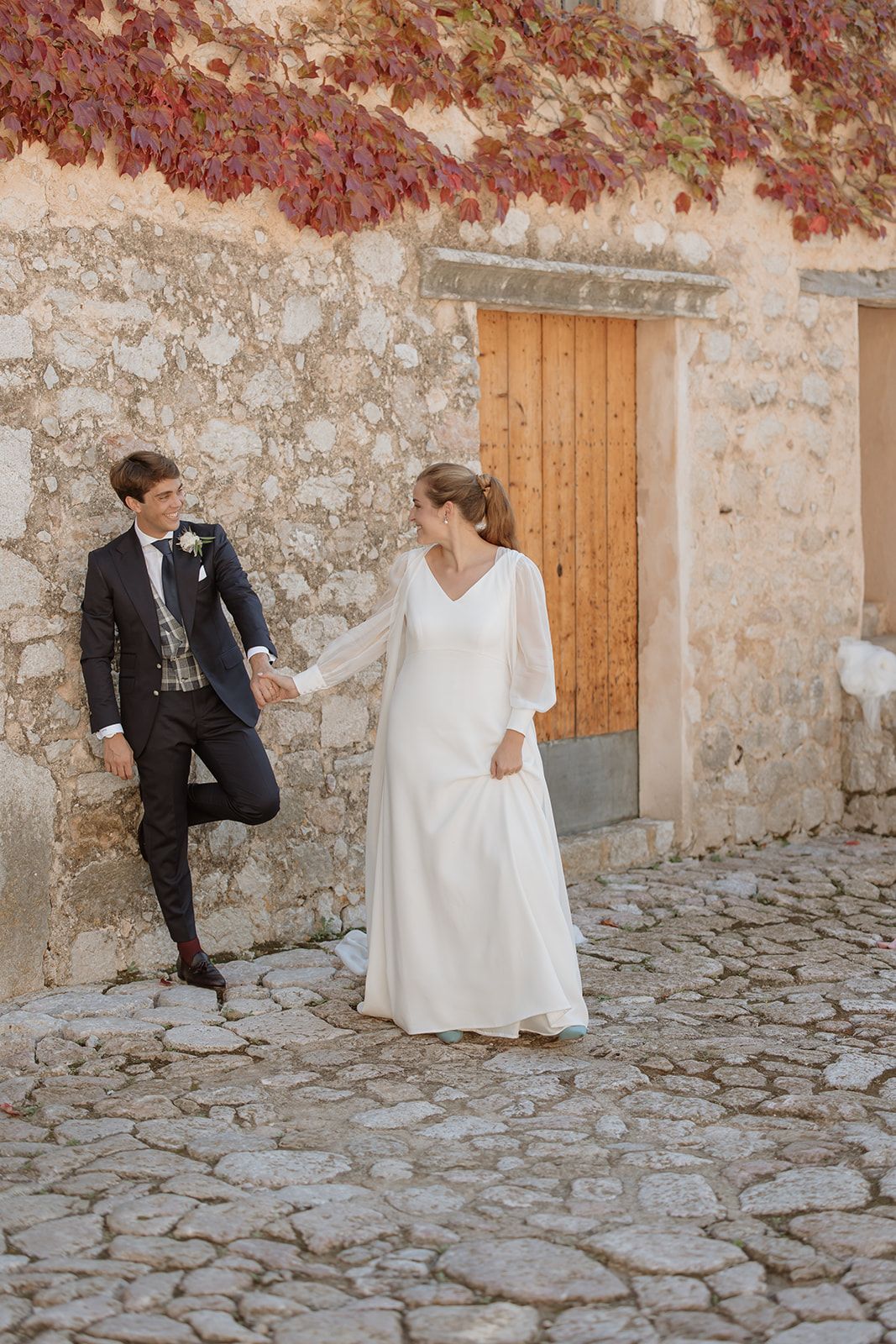 Wedding couple poses playfully by wooden door on stone wall with red autumn leaves.
