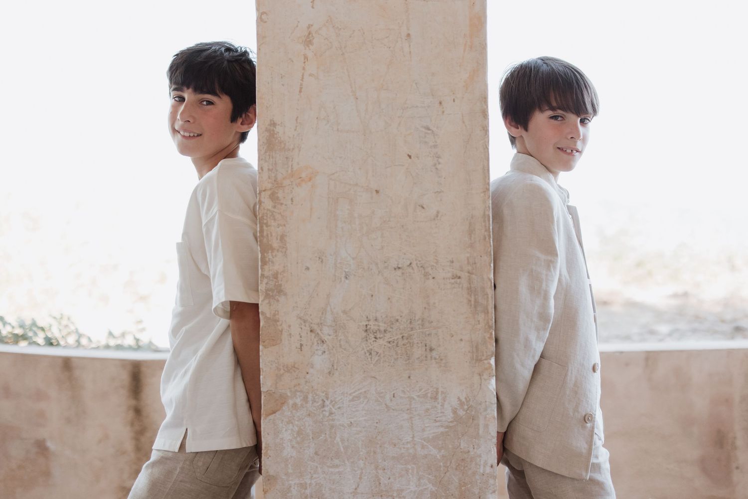 Two figures in white shirts stand back-to-back against a stone pillar in bright natural lighting.