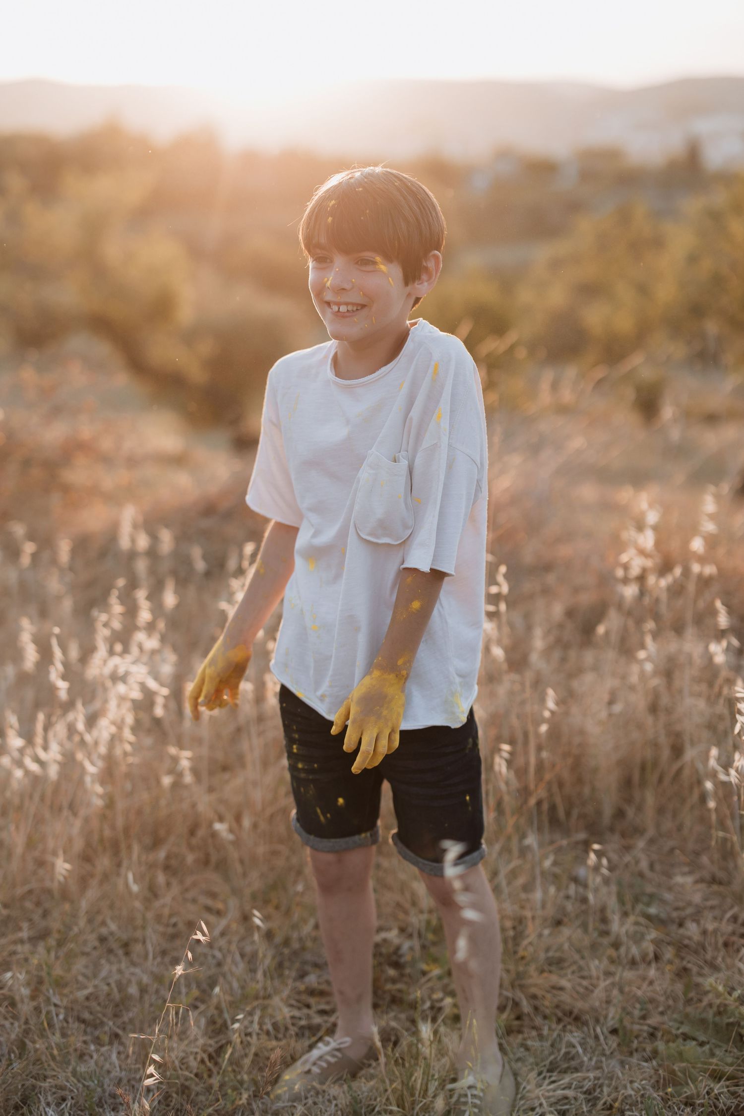 A child wearing a white t-shirt and yellow gloves stands in a sunlit meadow during golden hour.