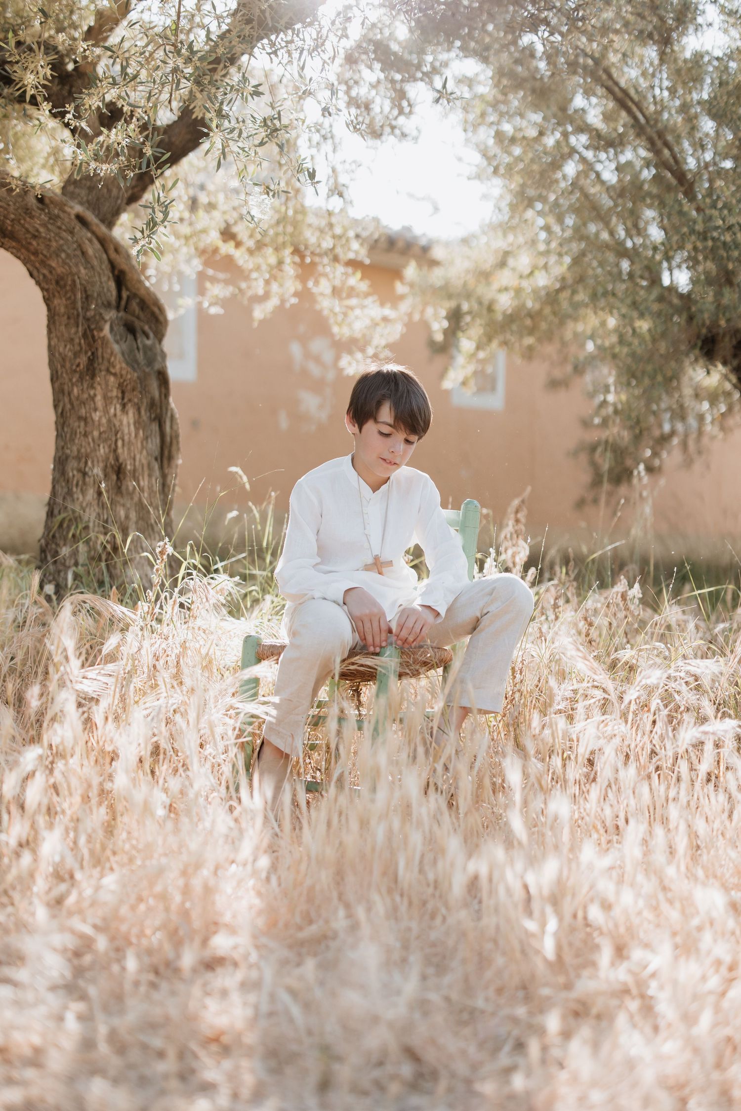 A figure in white clothing sits serenely among tall golden grass beneath a tree in warm sunlit afternoon.
