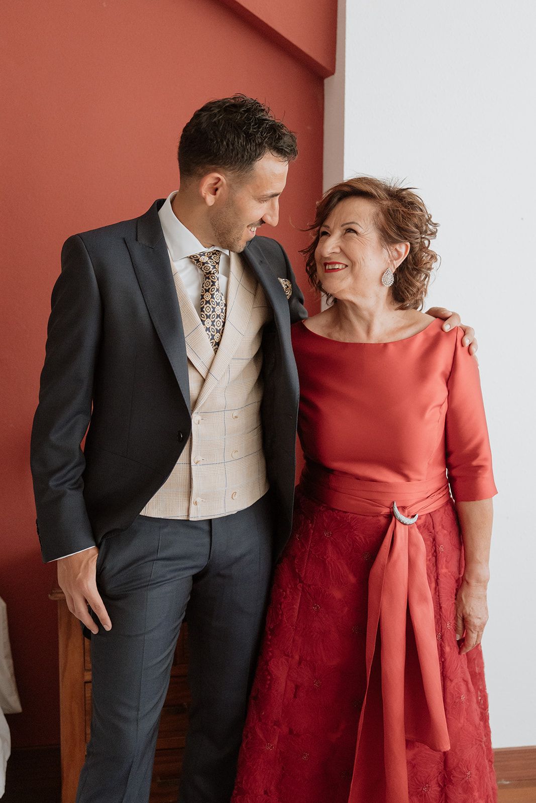 A formal couple stands against a coral wall sharing a joyful moment together.