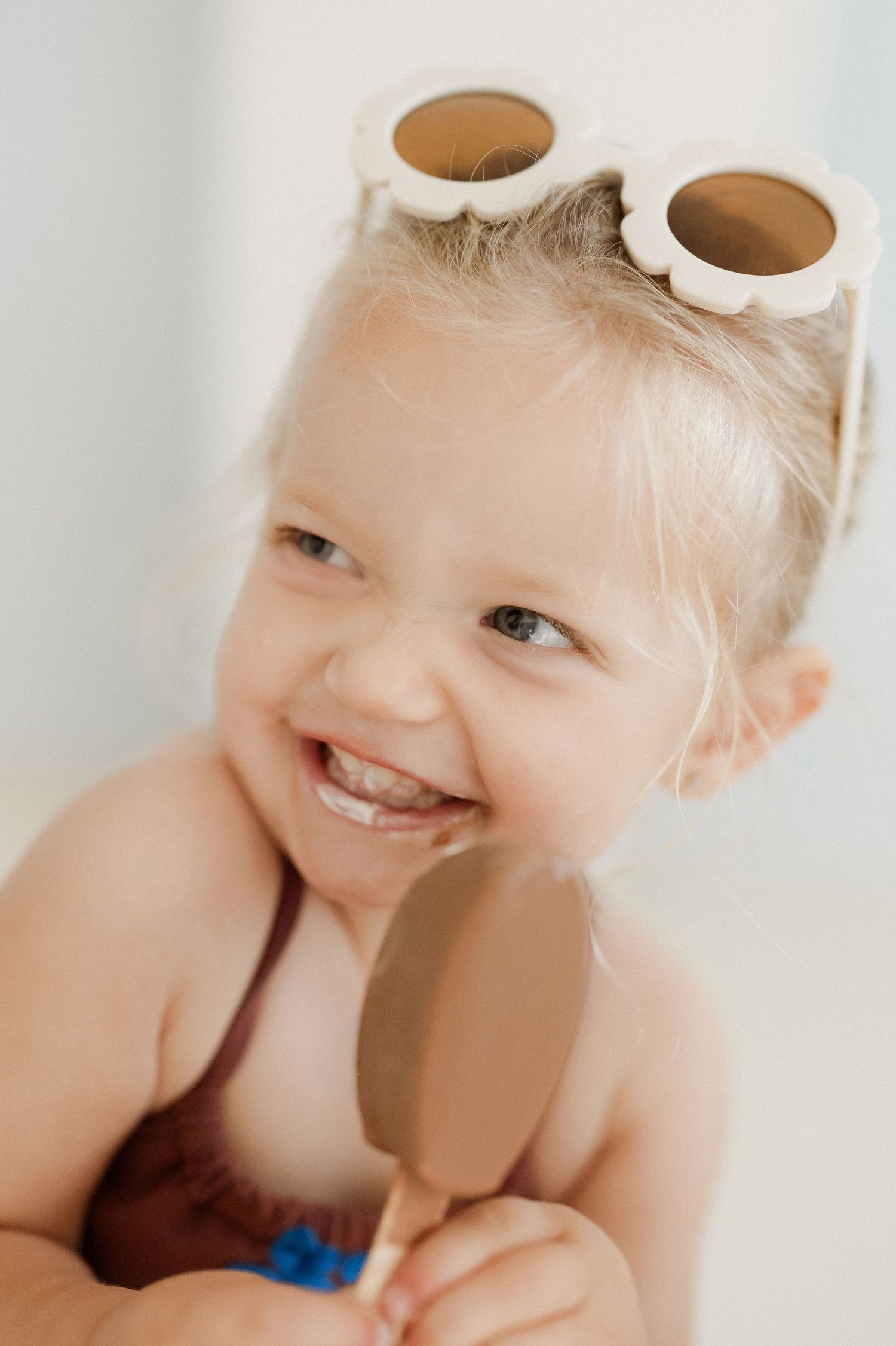 Happy toddler girl with sunglasses on head eating a chocolate ice cream bar in a swimsuit.