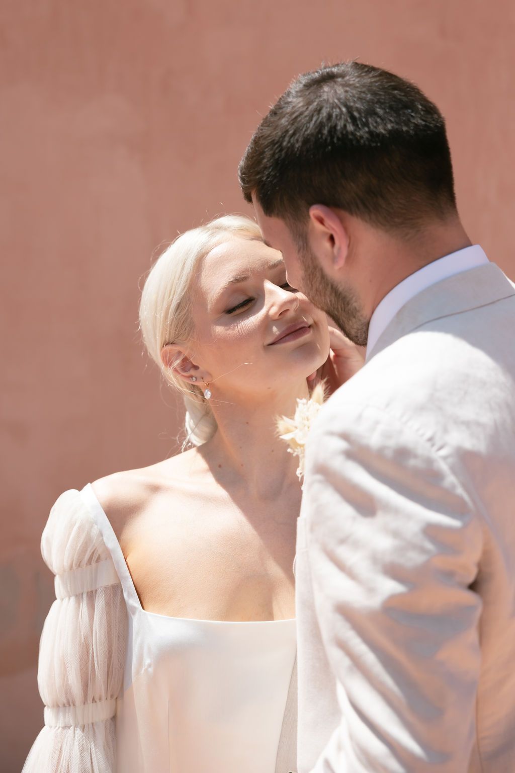 Intimate wedding portrait of a couple in white attire against a pink wall.