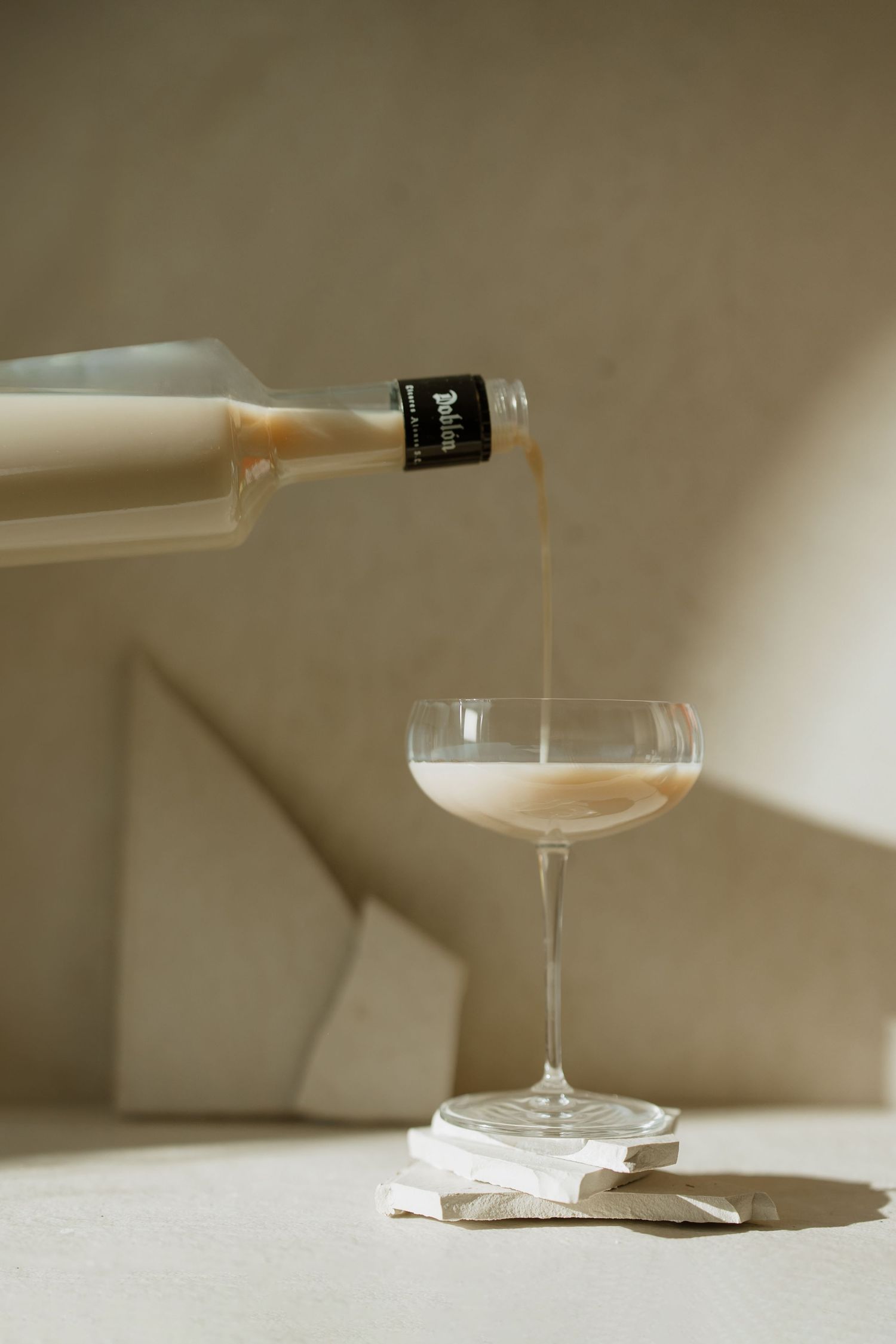 Creamy liquid being poured from a bottle into an elegant coupe glass on a white surface with soft shadows.