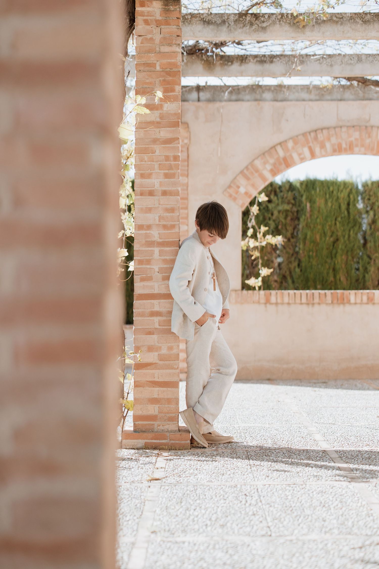 Person in all-beige outfit leans against brick column under Mediterranean-style archway at outdoor venue.