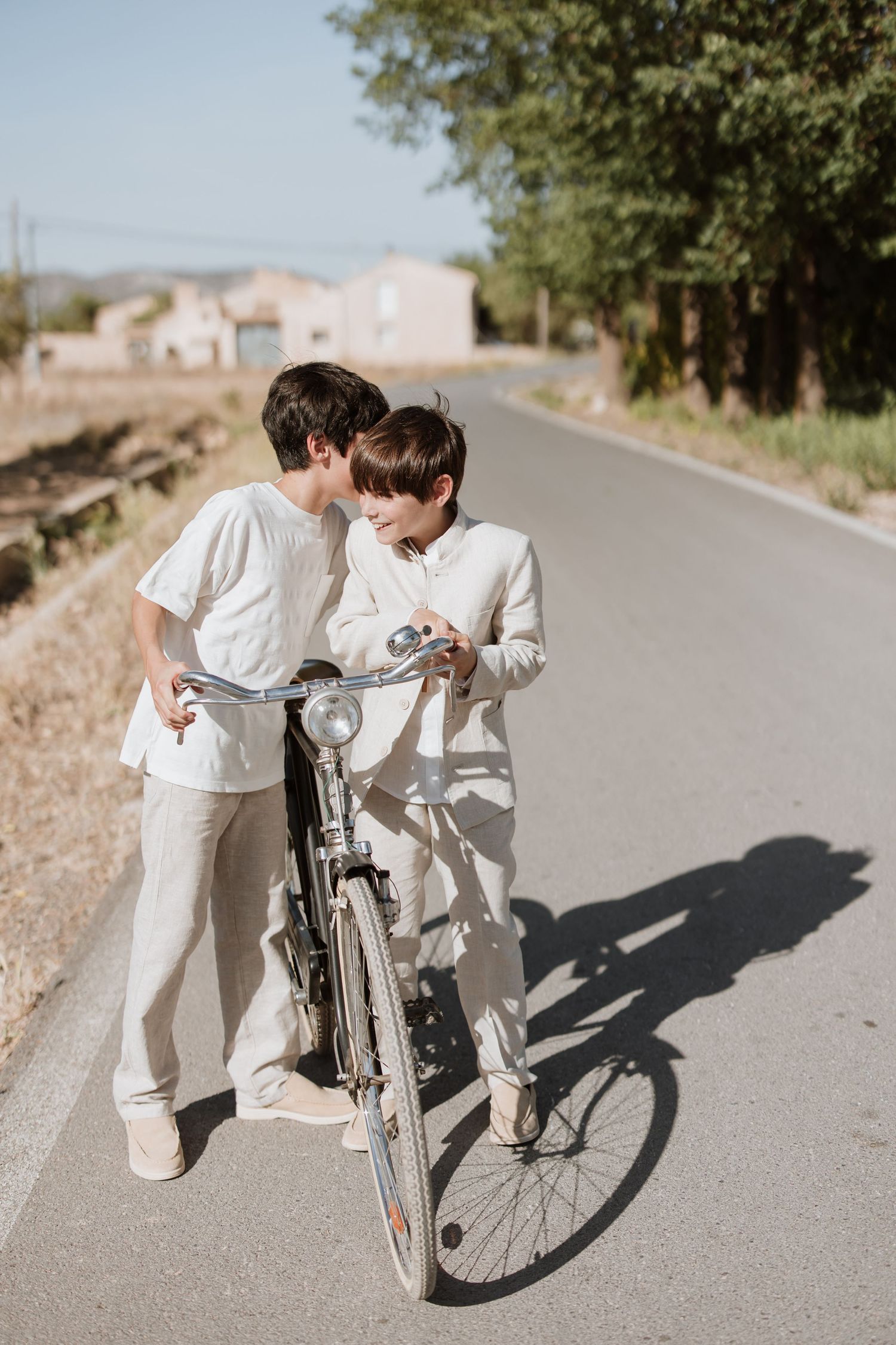 Two people in white clothing standing with a vintage bicycle on a sunny country road with houses in background.