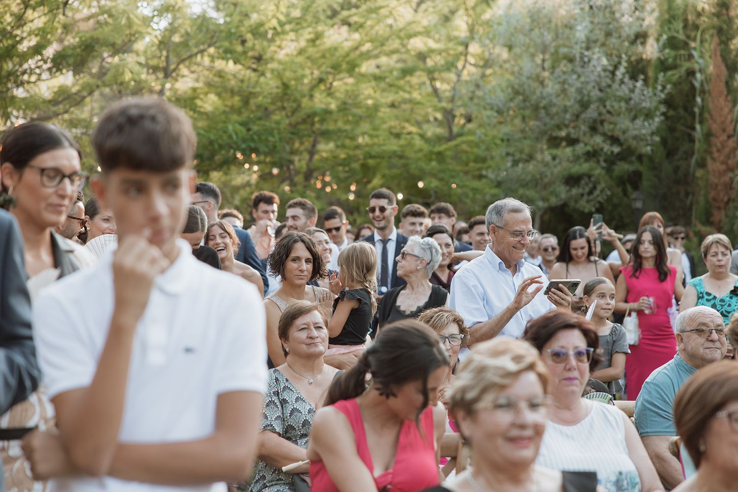 A crowd of wedding guests seated outdoors watching a ceremony in a natural setting.