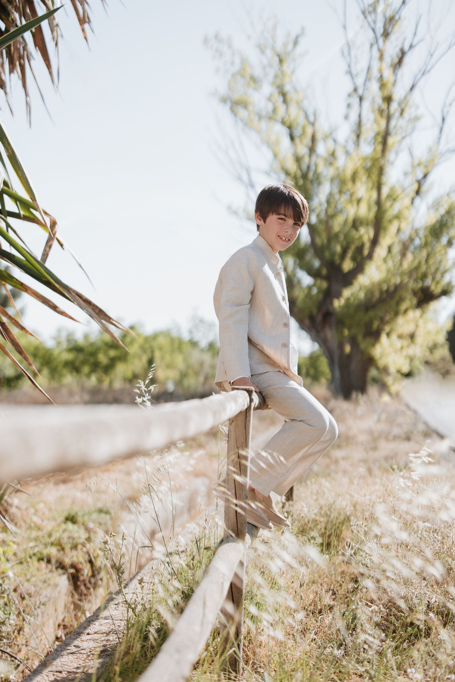 Young person in light clothing sitting and smiling in a sun-drenched field of tall grass on a bright day.