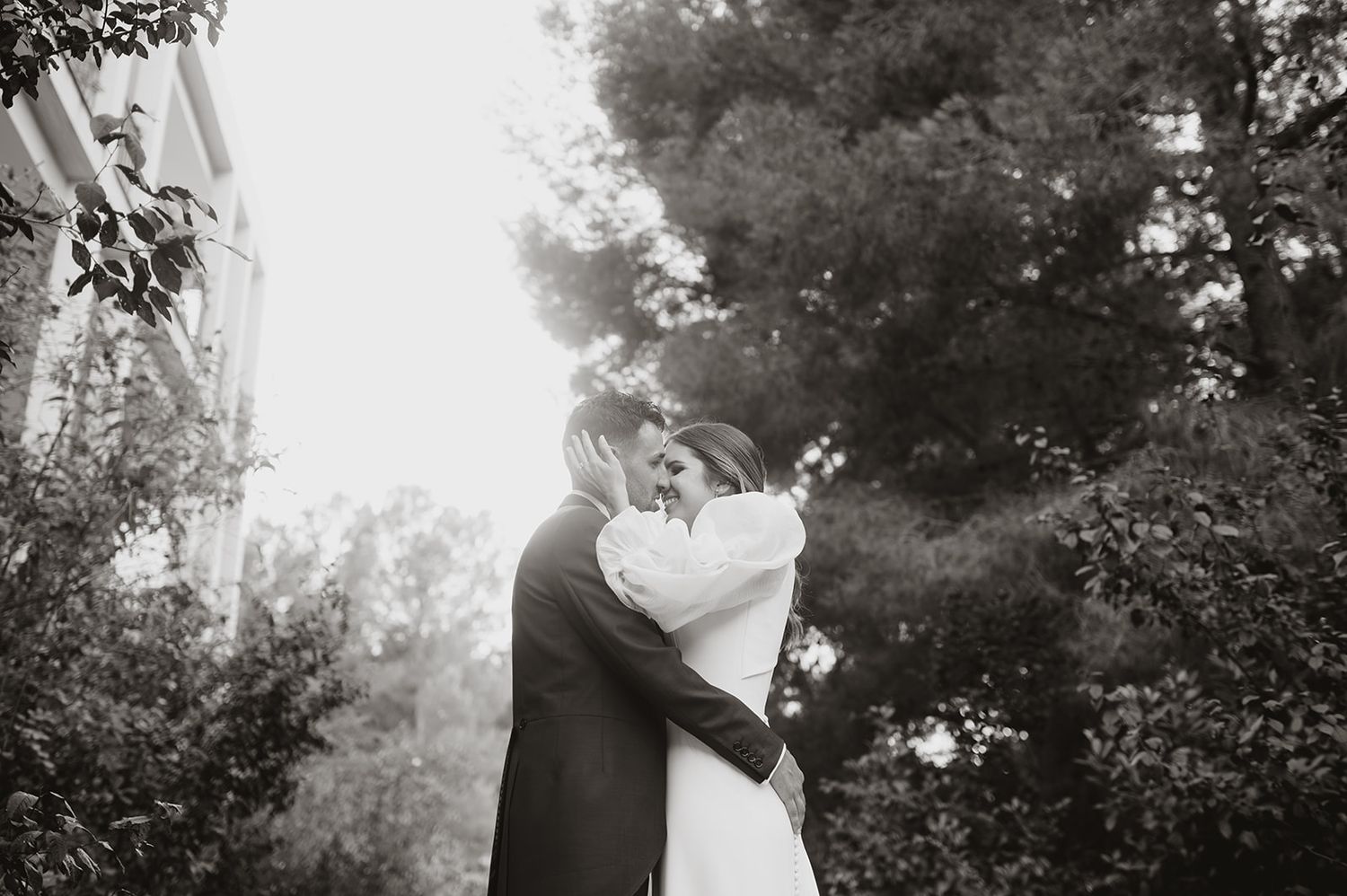 Black and white wedding photo of couple embracing outdoors with sunlight filtering through trees.