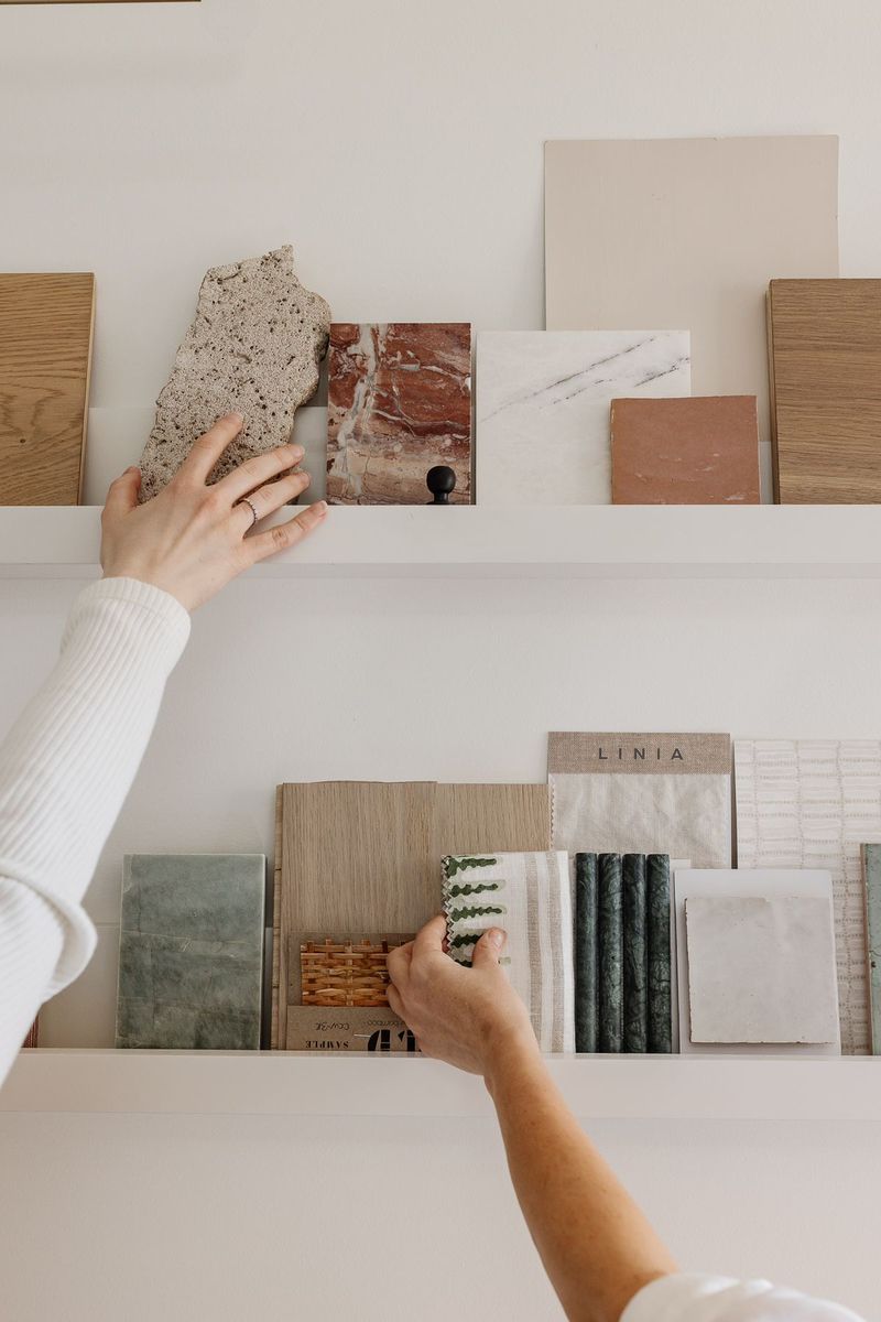 Person selecting material samples from white shelves displaying tiles, wood, marble, and stone swatches in a design studio.