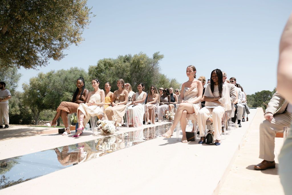 Wedding guests seated on outdoor benches in light colored attire.