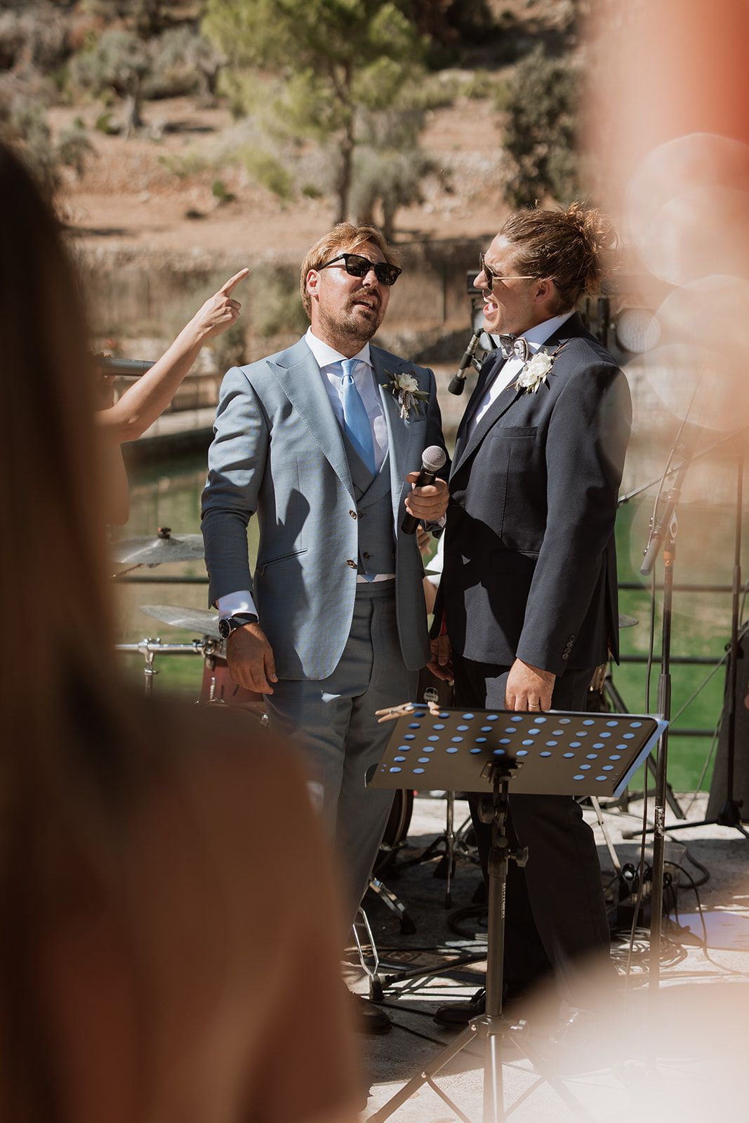Two performers in suits stand at a wedding ceremony podium outdoors with music stands and equipment.