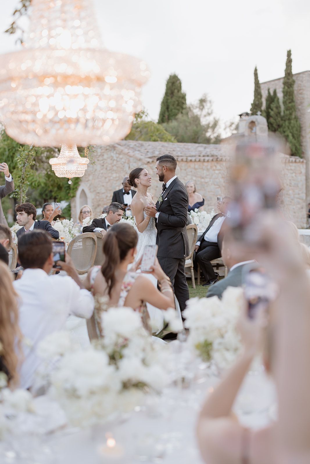 Outdoor wedding reception with chandelier and white floral decorations at dusk.