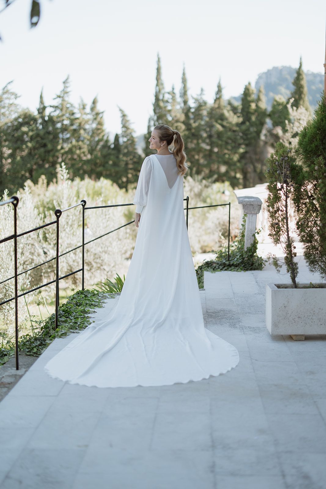 Bride in flowing cathedral-length cape standing on terrace with mountain views.