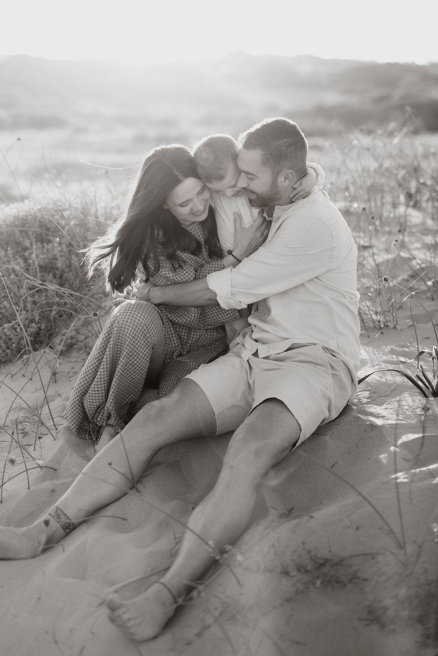 Black and white photo of a happy family sitting on sandy beach dunes, parents embracing their child warmly.