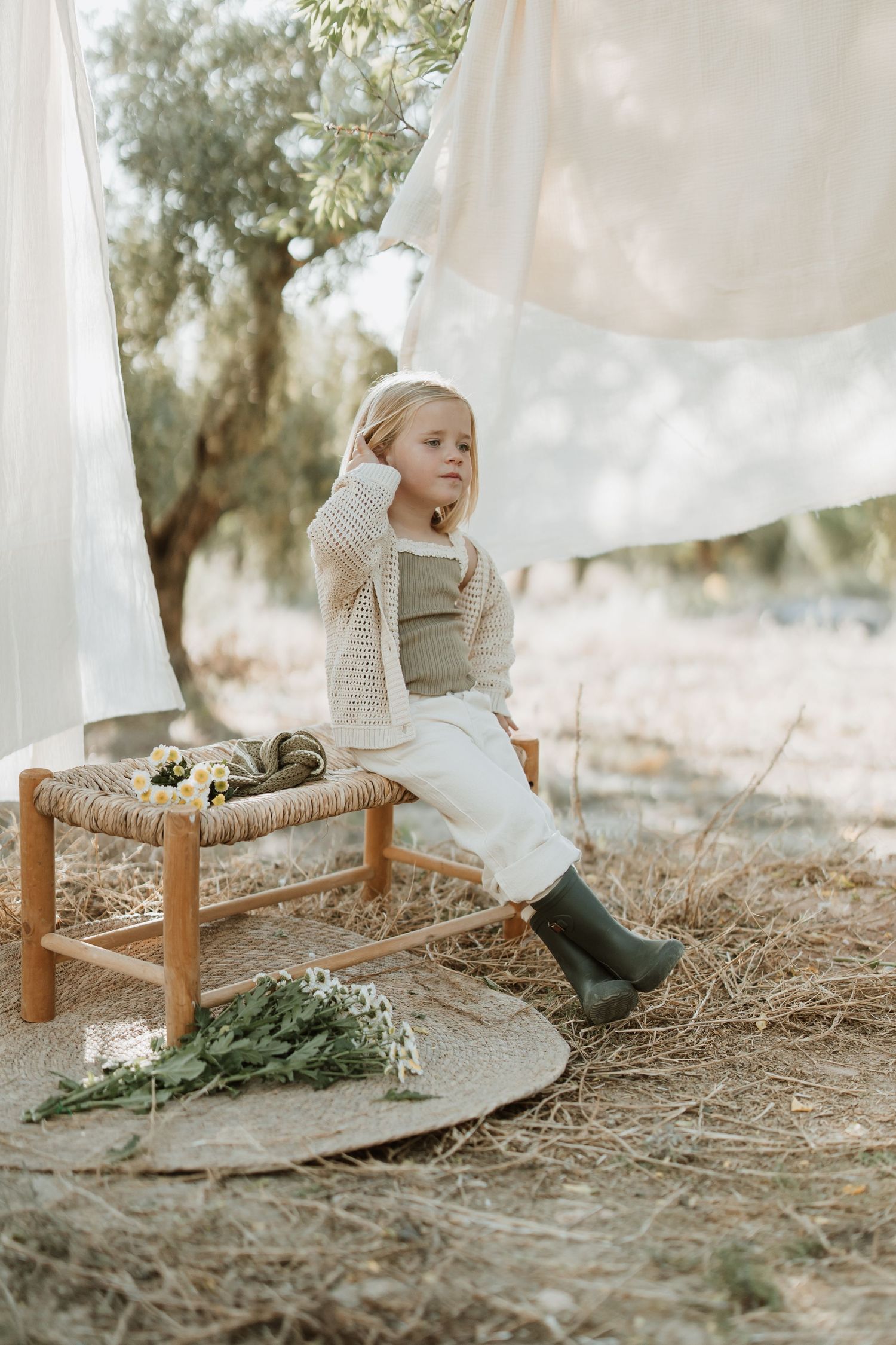 Figure in cozy sweater and boots sits on rustic bench among hanging white linens.