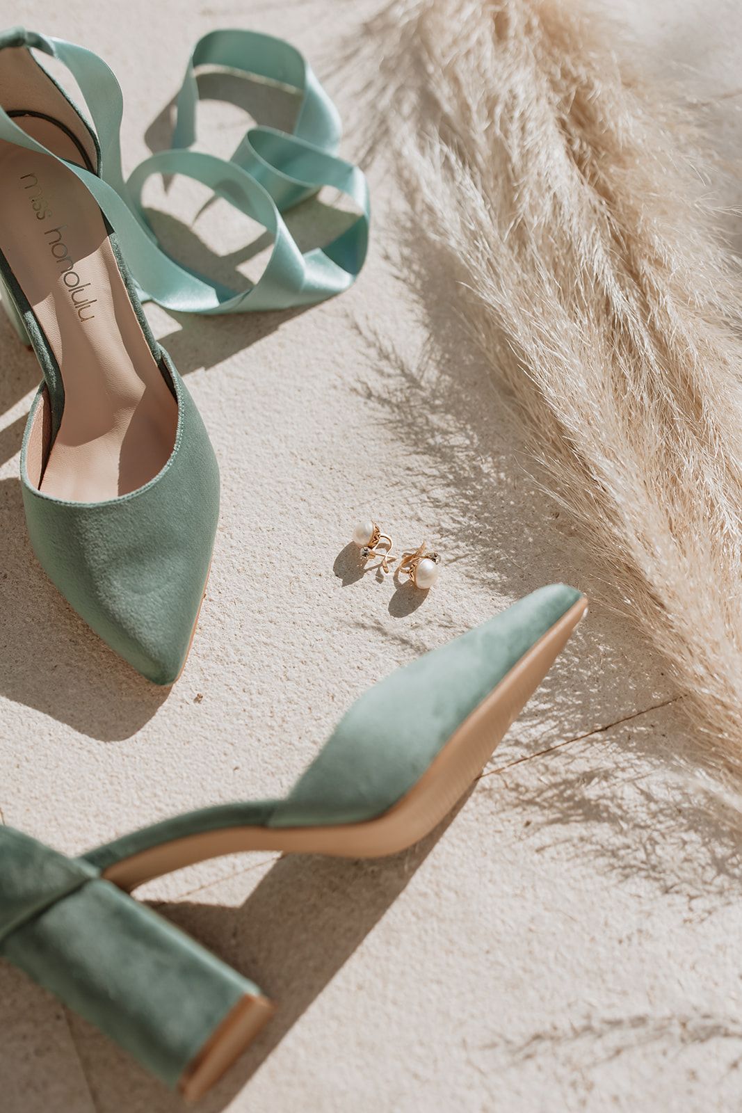 Mint green wedding shoes and earrings styled with pampas grass on white surface.
