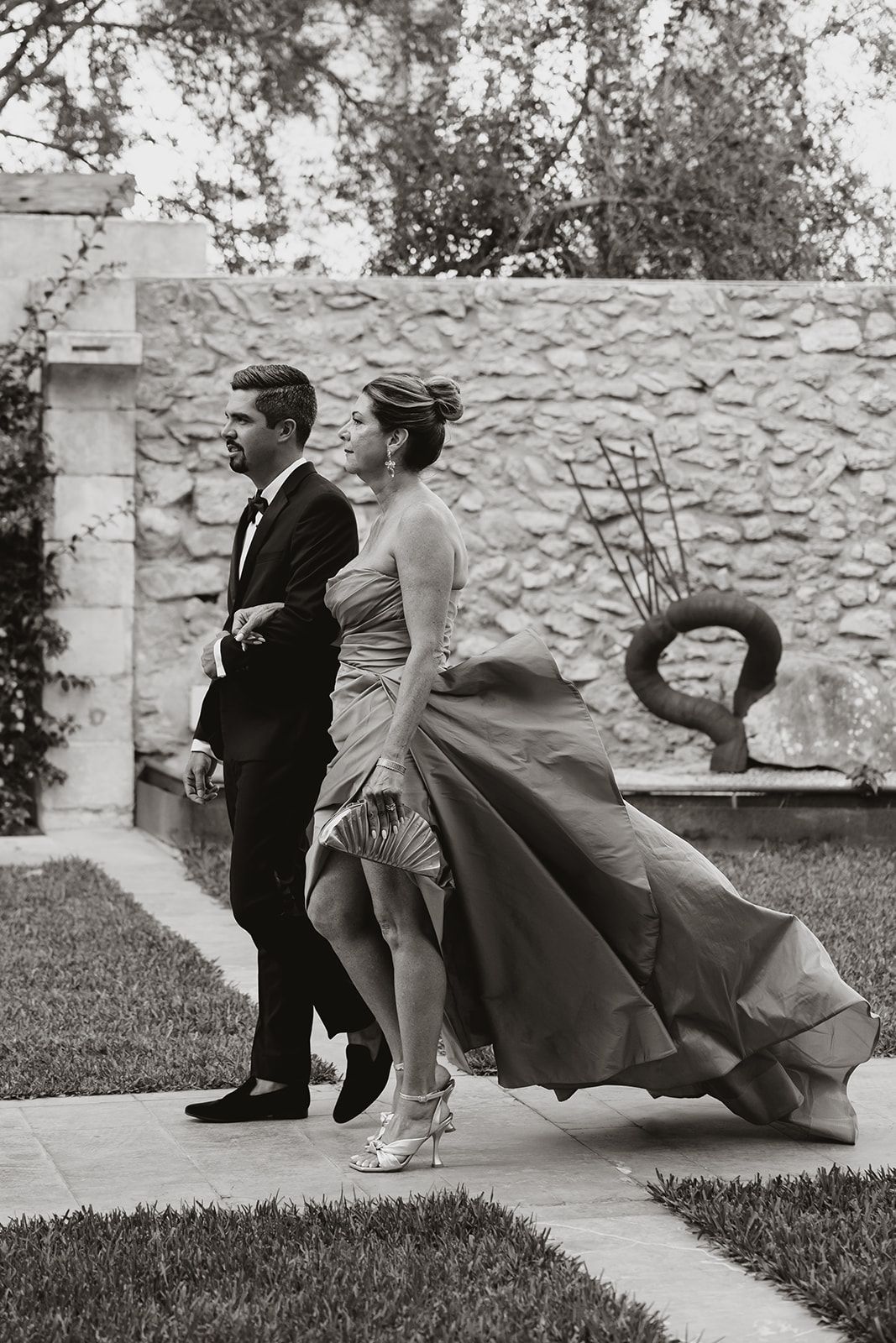 Black and white photo of a couple in formal attire walking on a garden path beside a stone wall.