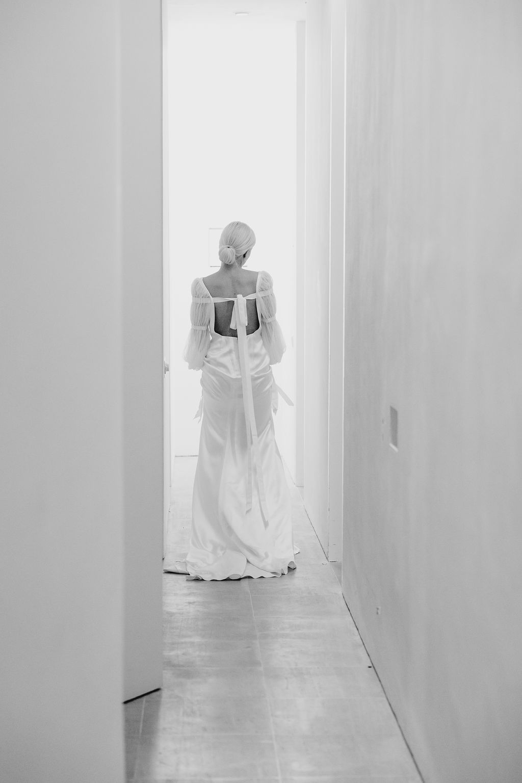 A black and white photograph of a wedding dress captured from behind in a narrow hallway.