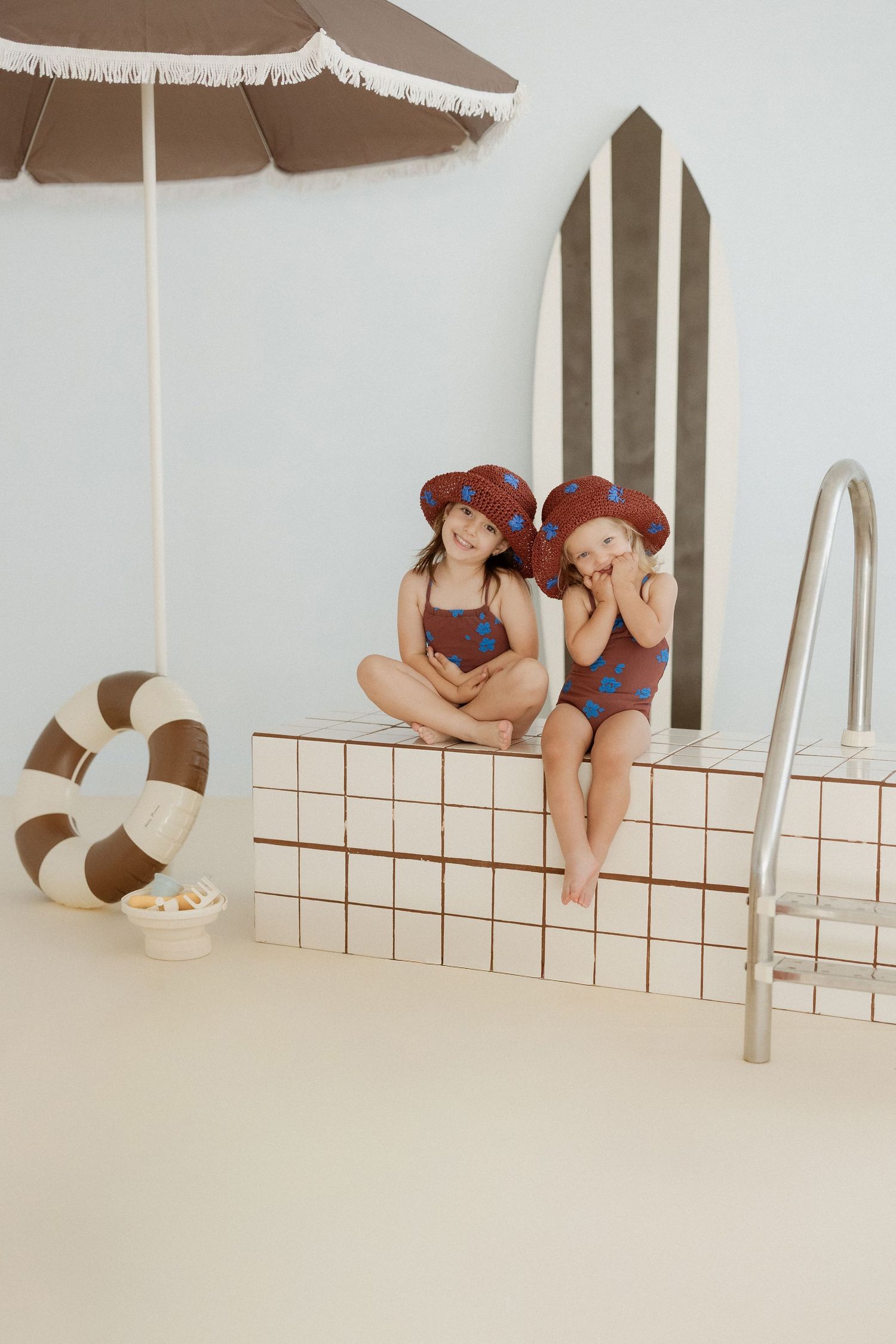 Two young girls in matching floral swimsuits and hats sit poolside near a striped surfboard and beach umbrella.