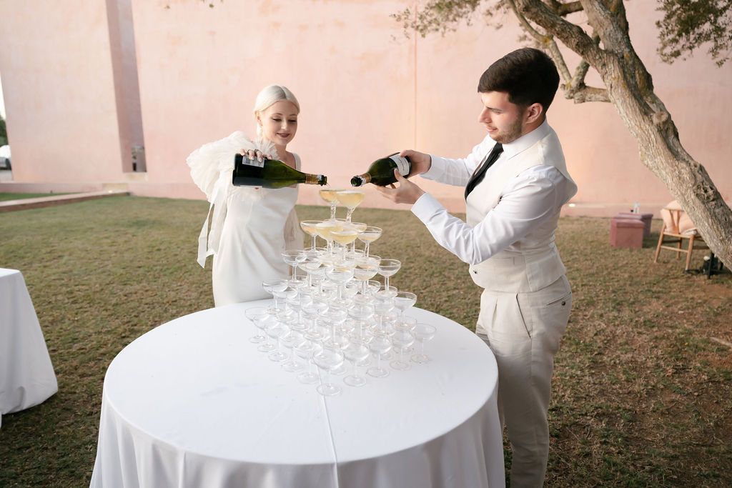 Couple pouring champagne into tower of glasses at outdoor wedding reception.
