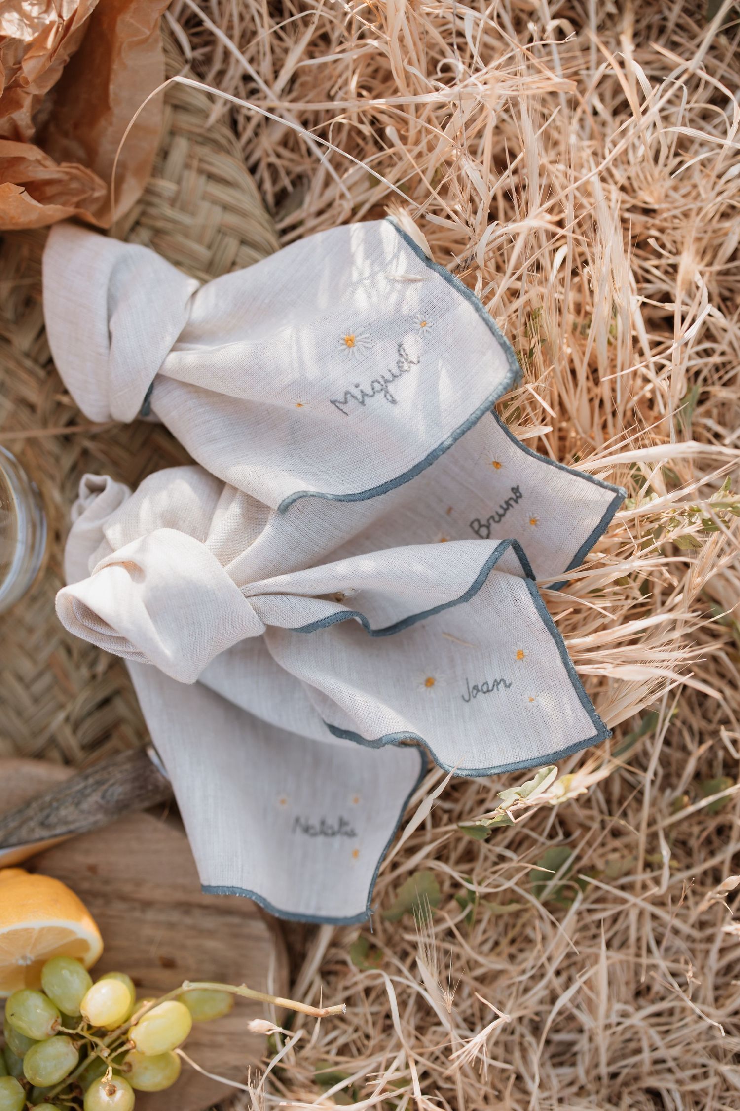 White linen napkins with delicate embroidery details arranged on straw with grapes and citrus fruits in a rustic setting.