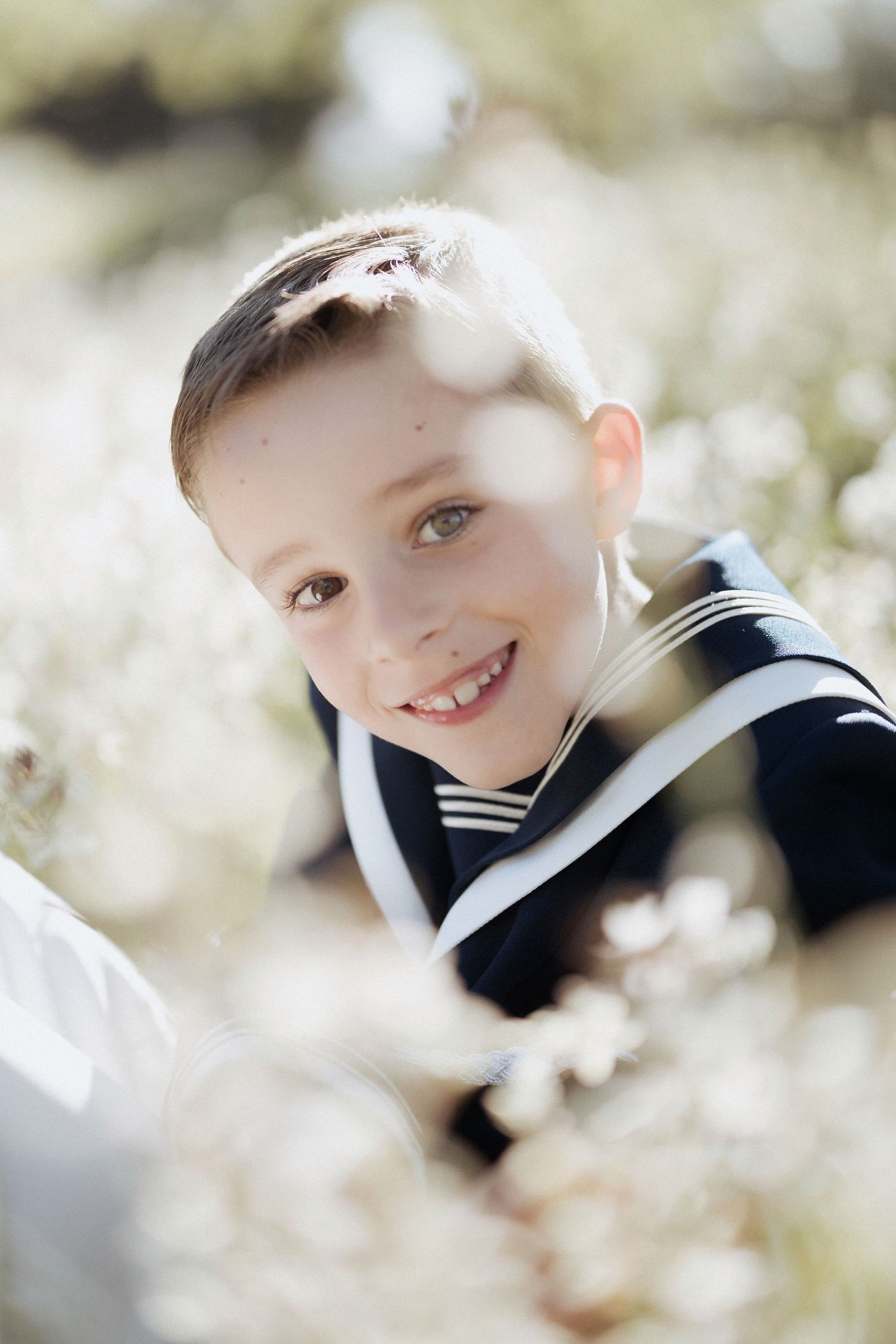 Smiling boy in navy sailor outfit surrounded by white wildflowers in a sunny outdoor setting.