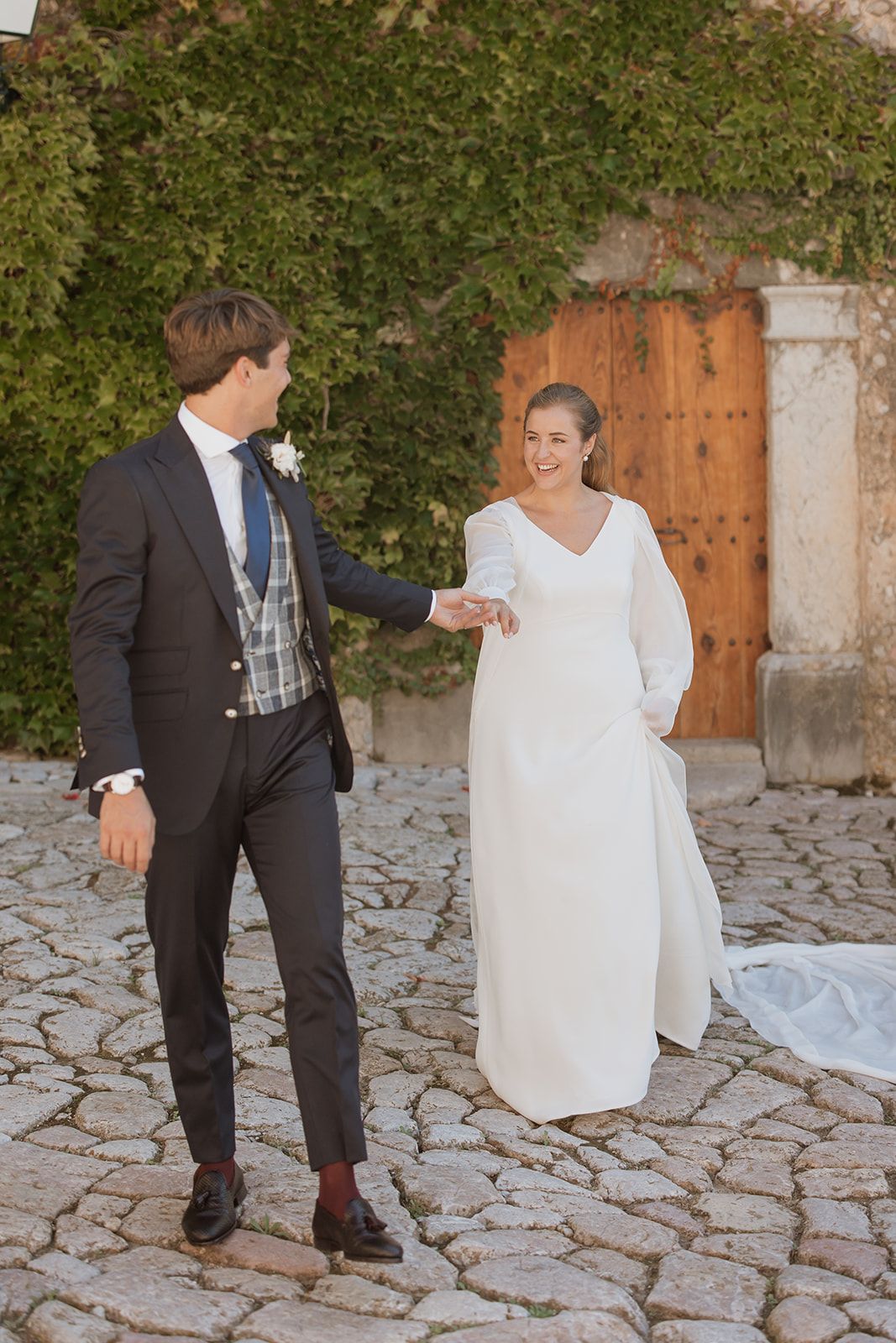 Bride and groom share a joyful moment on cobblestone path by rustic wooden door.