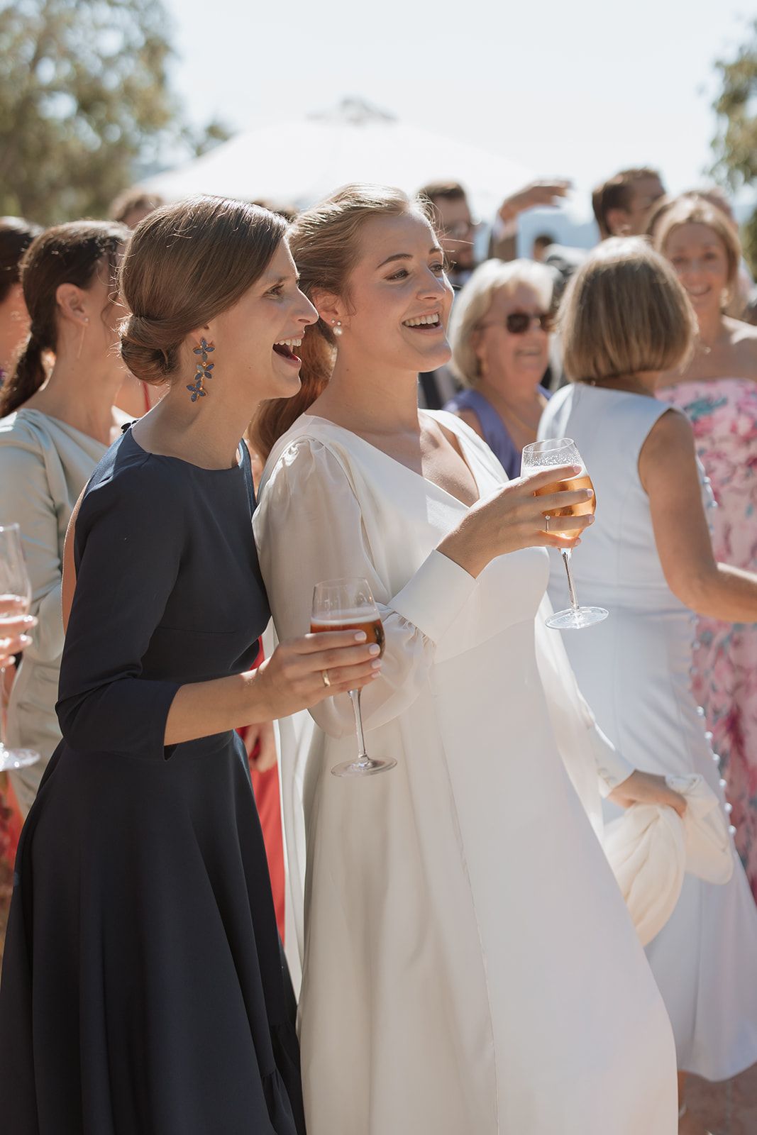 Wedding guests in formal attire smile while holding champagne glasses at an outdoor celebration.