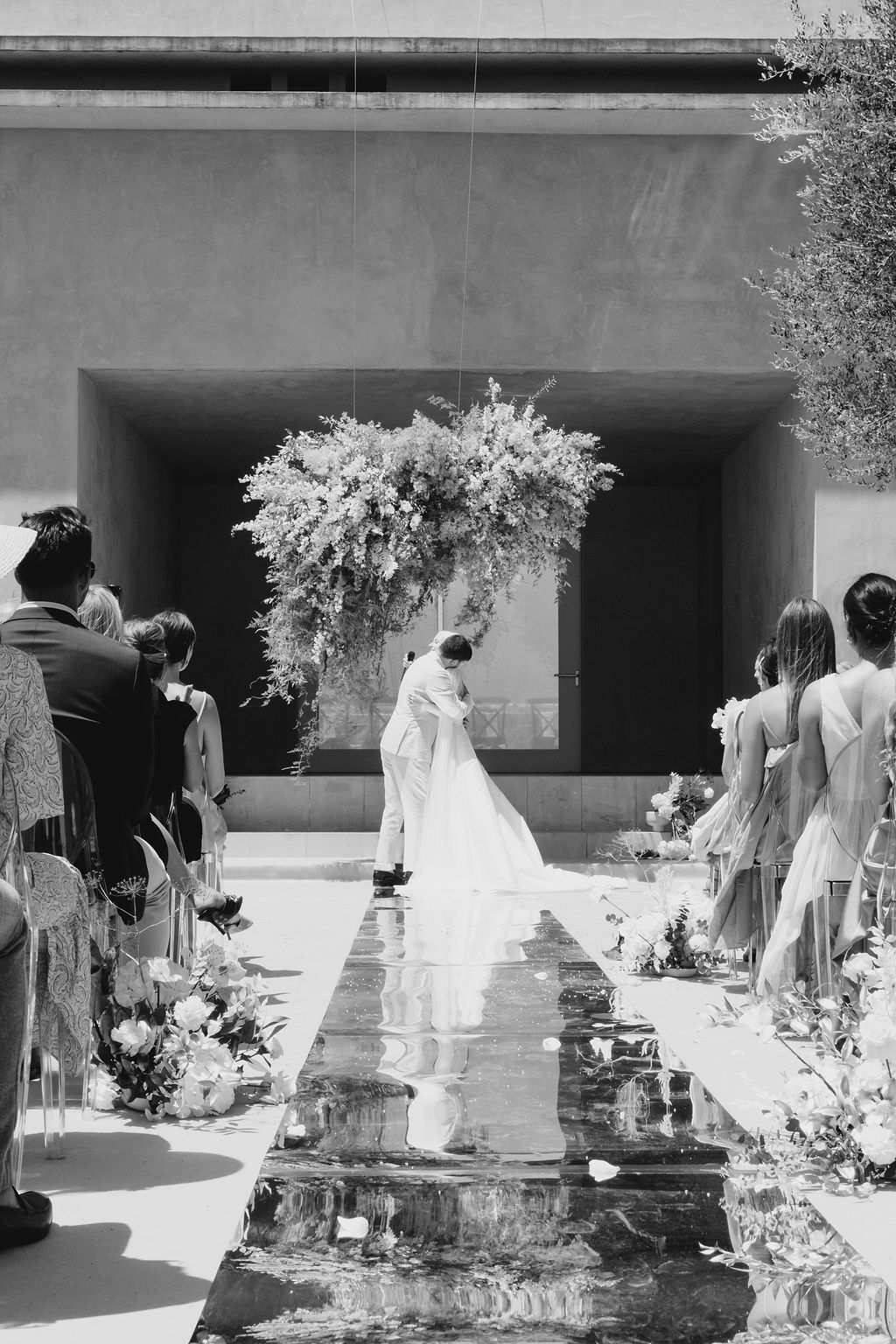 Black and white photo of a wedding ceremony with a reflective aisle and floral arch.