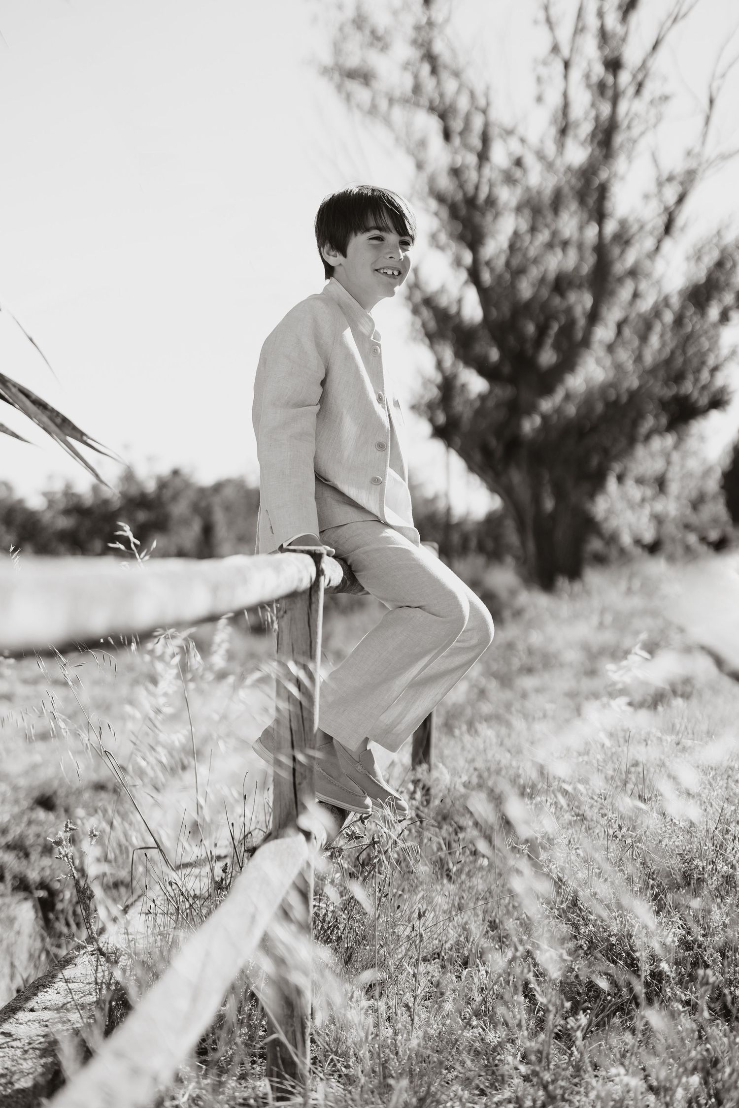 A person in a light-colored suit sits casually on a rustic fence in a monochrome outdoor setting with trees in the background.