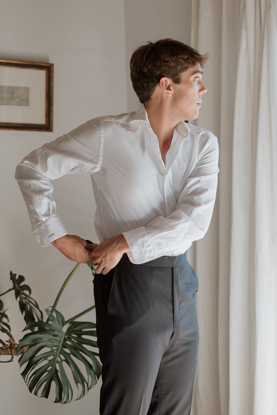 Side profile of person in white dress shirt adjusting sleeves near window with monstera plant.