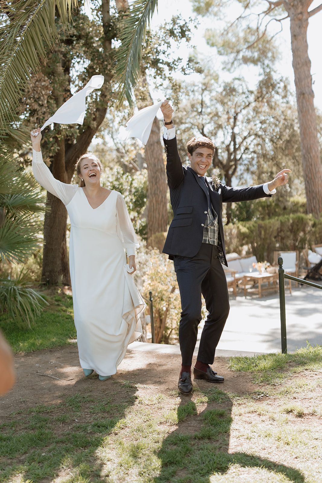 Newlyweds celebrate with raised arms and waving napkins during outdoor garden celebration.