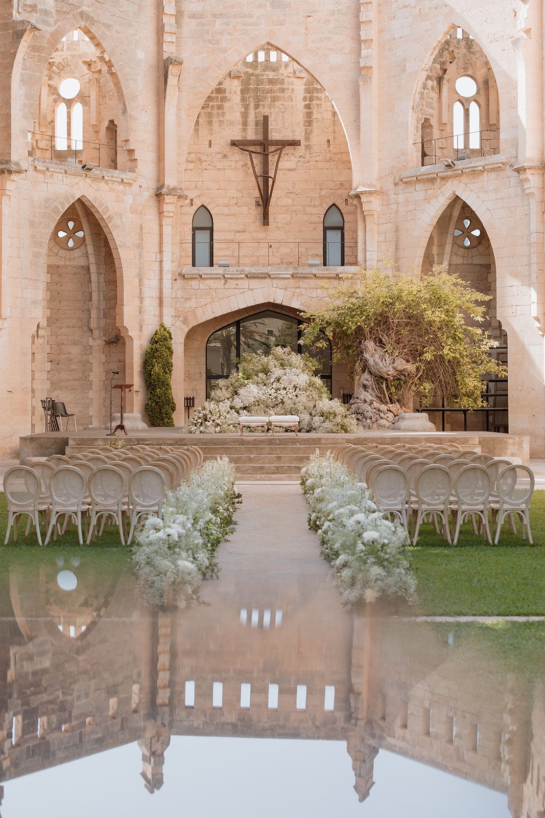 Religious ceremony venue with white chairs and floral decor reflected in water feature foreground.