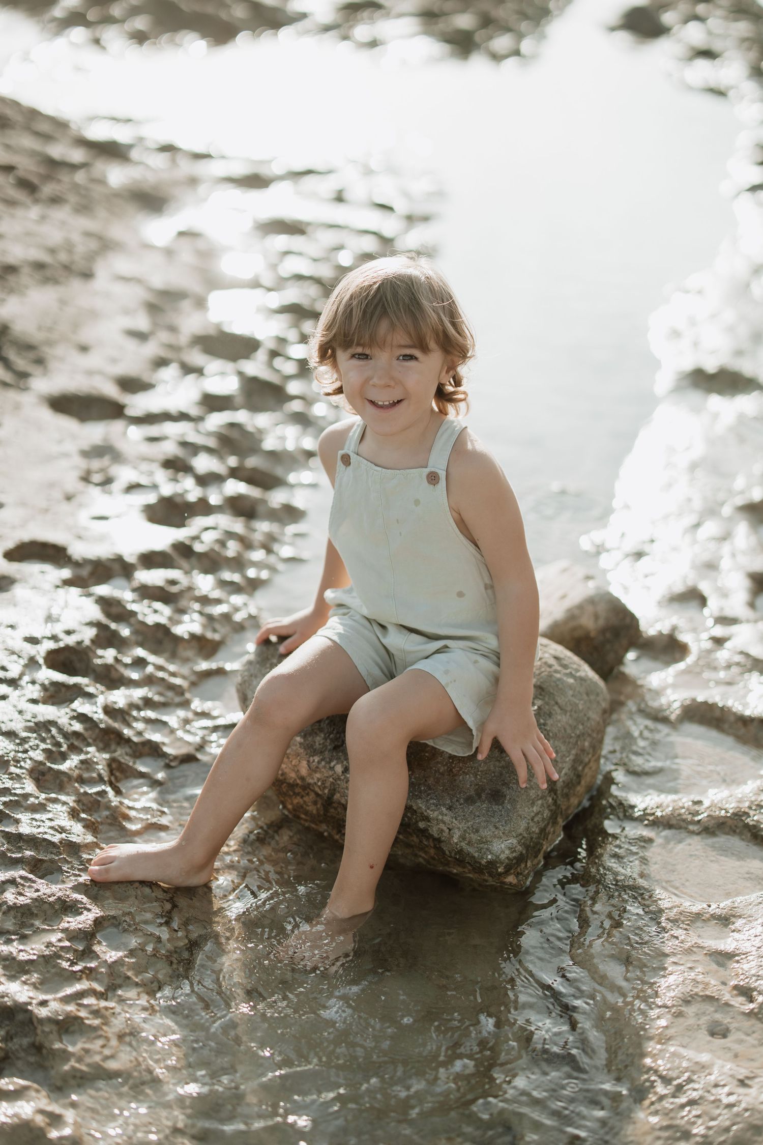 Child in light colored clothing smiles while sitting on beach rock during sunset.