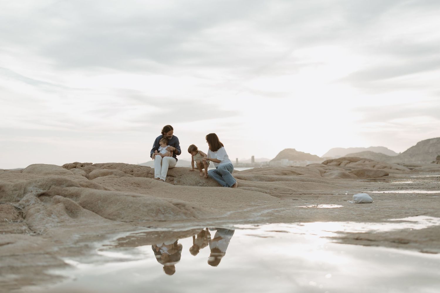 Group sitting on rocky shoreline with mountains in background and reflection in water.