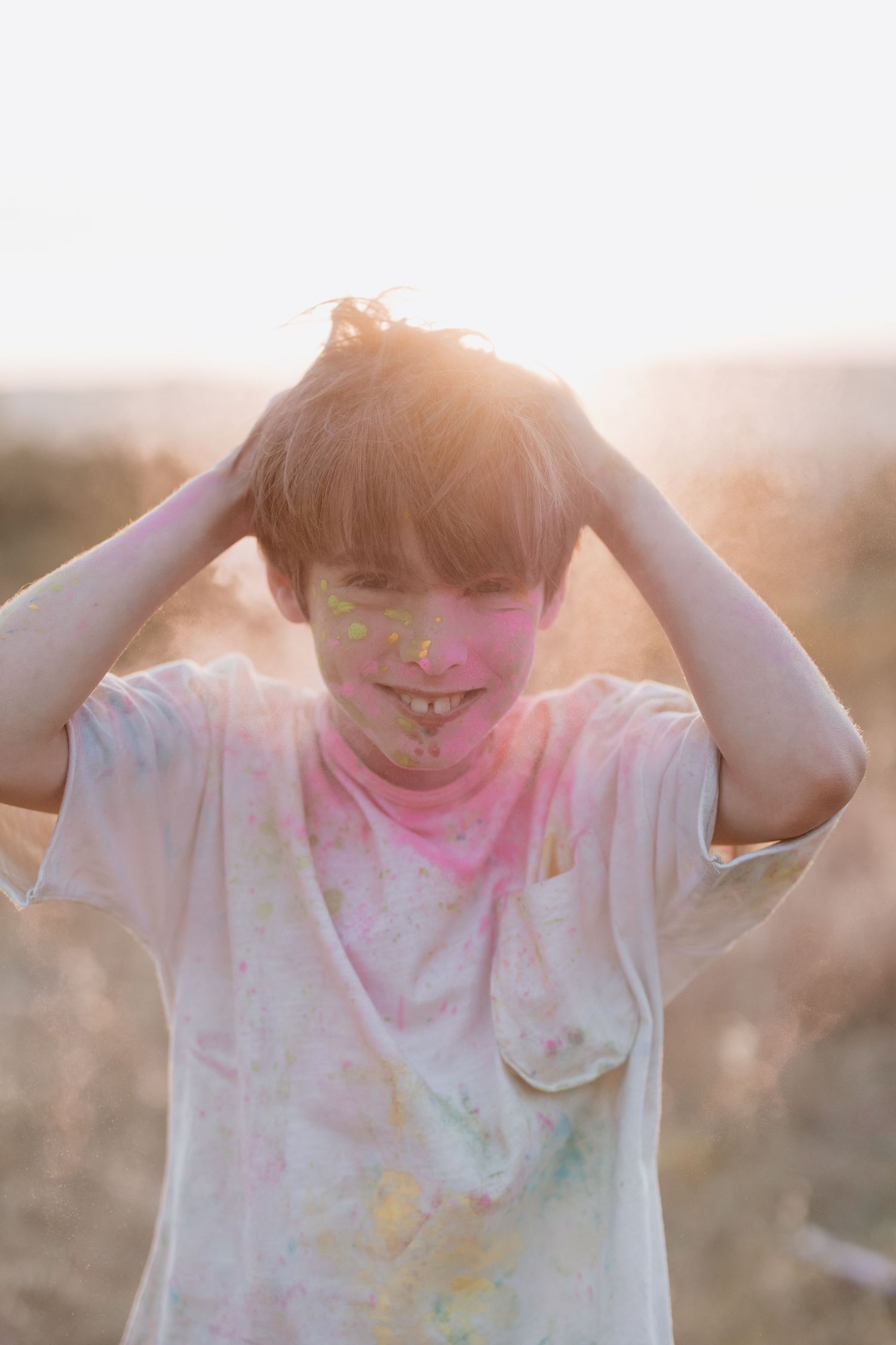 Person covered in colorful powder smiles while playing with their hair during golden hour sunset outdoors.